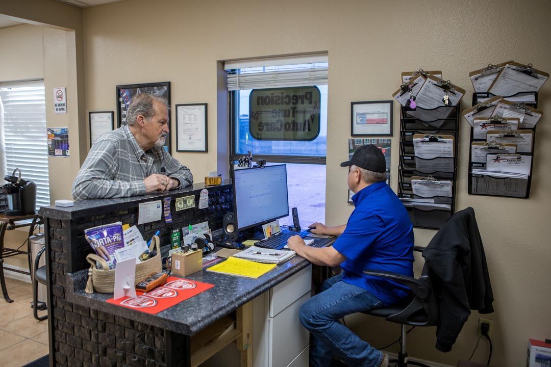 Two men talking at a cluttered office reception desk. One man stands; the other sits at a computer.