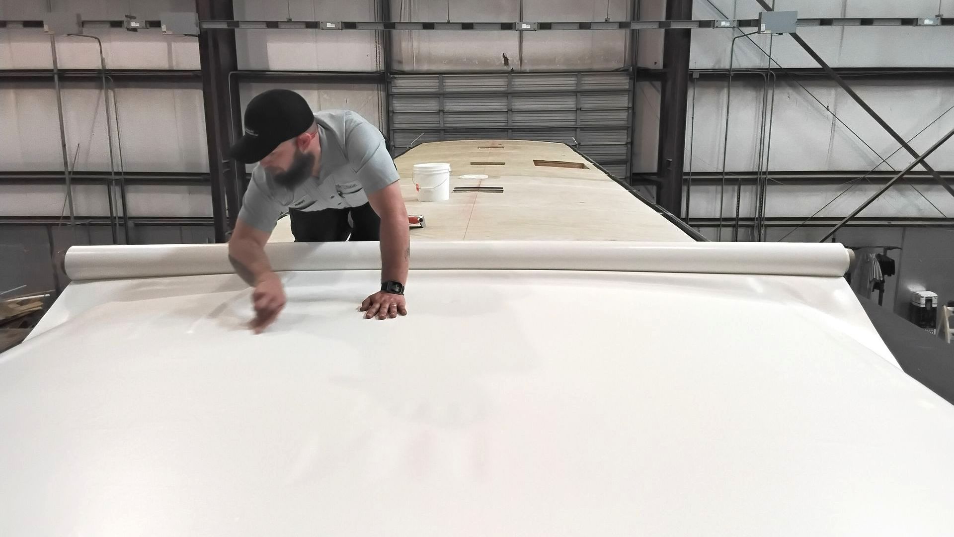 A person in a gray shirt rolls out a large white sheet of roofing material across an RV roof in a warehouse.
