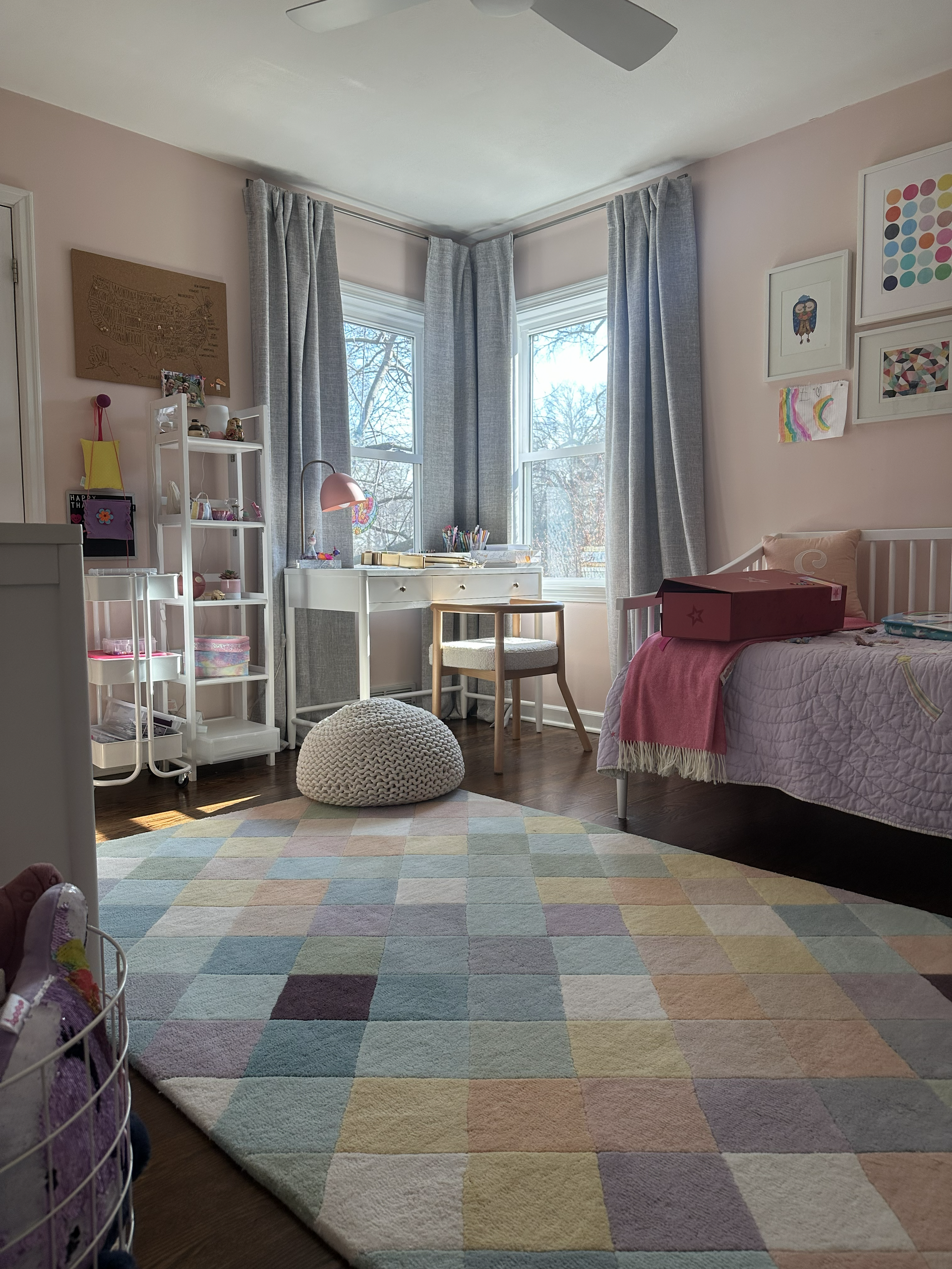 Small, cozy bedroom with a colorful rug, bay window desk, bed, and shelving in soft pastel tones.