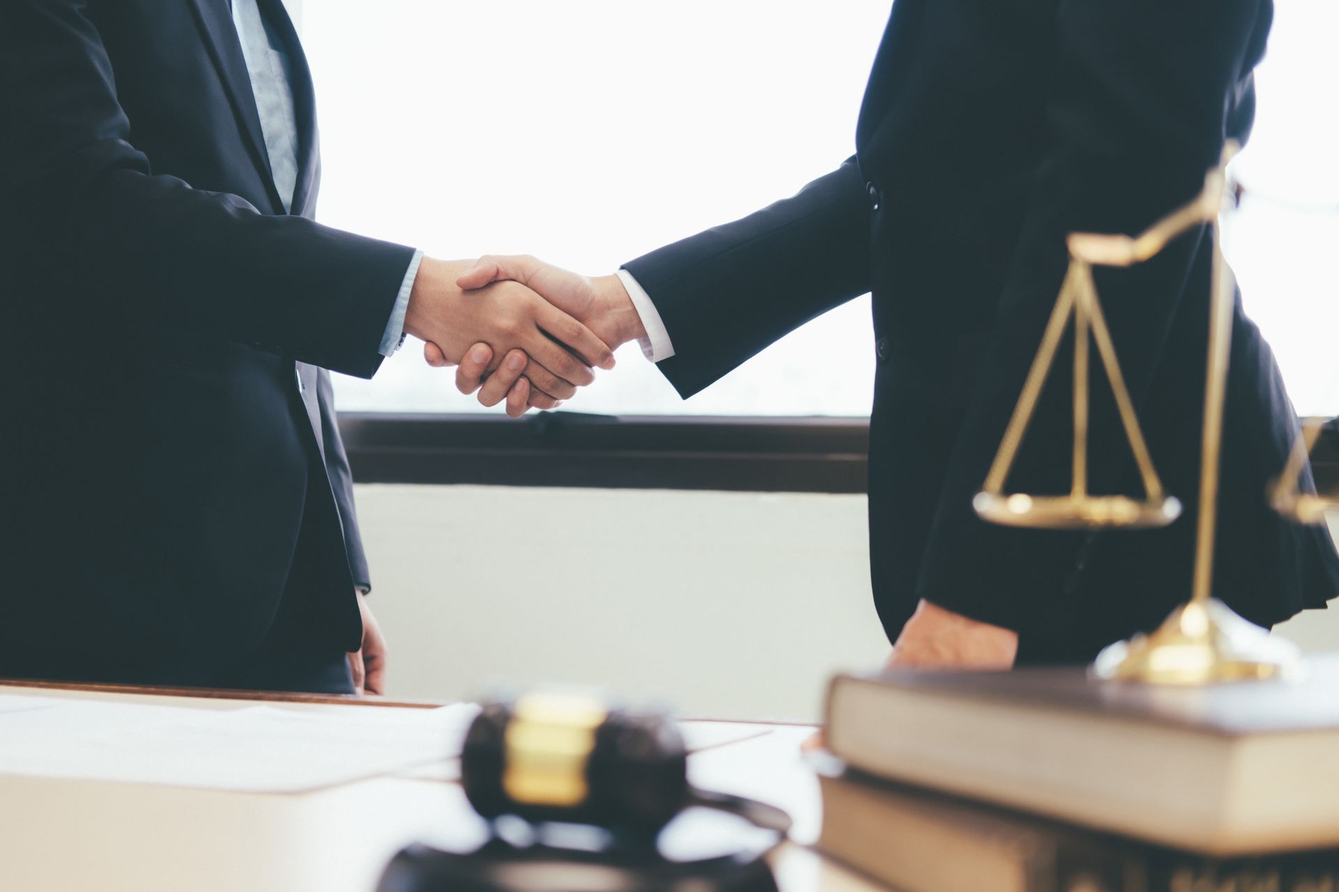 Two people in suits shaking hands in an office, gavel and scales of justice in the foreground.
