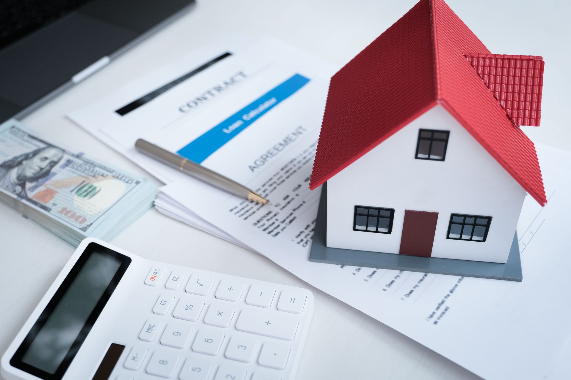 Miniature house, documents, calculator, money, and pen on a table, representing real estate.