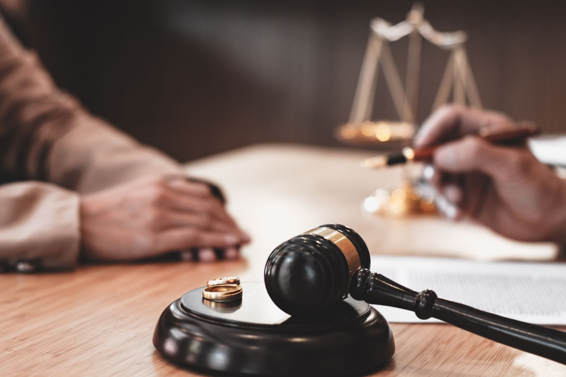 Wedding rings, gavel, and scales of justice on a table with people, likely in a legal setting.