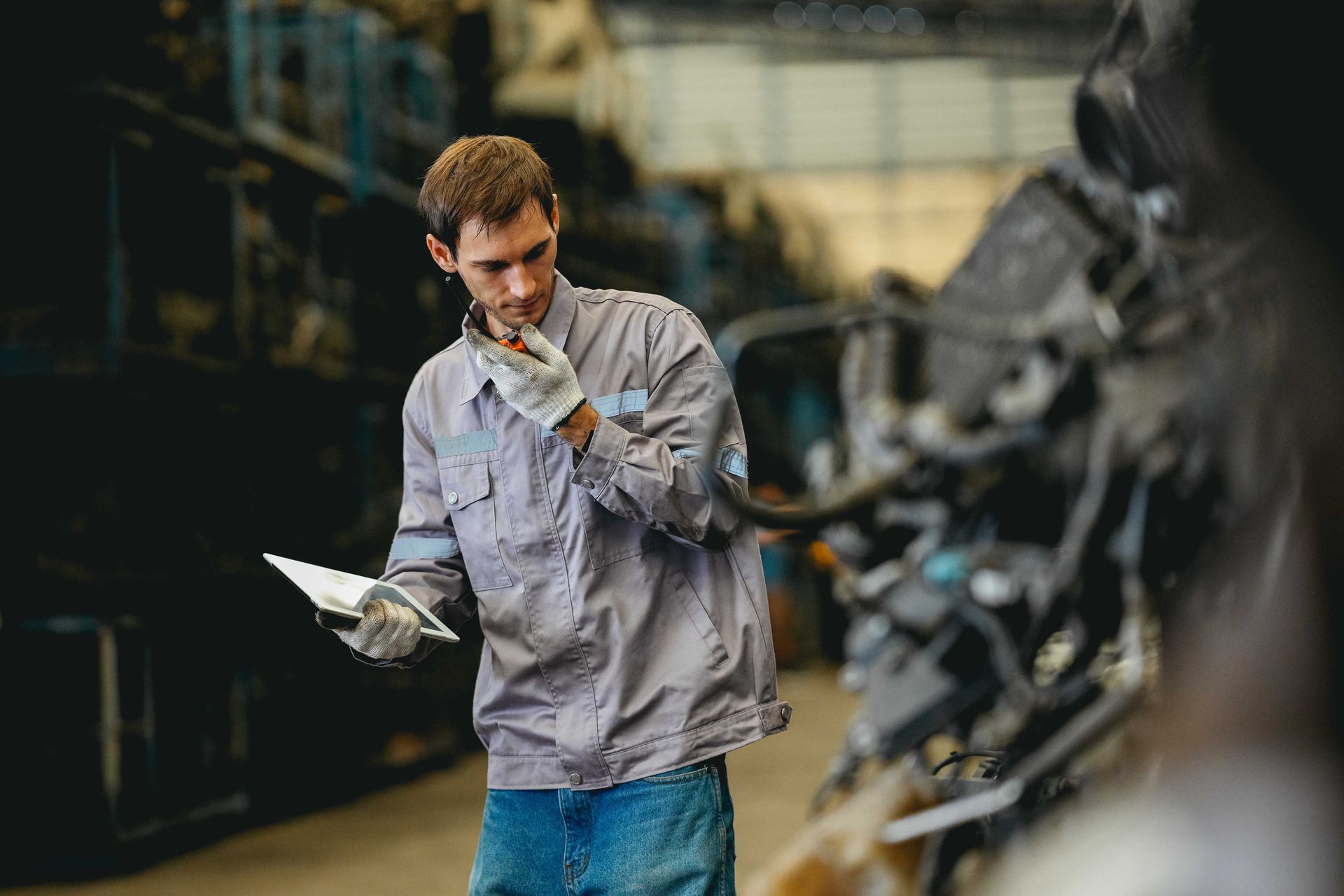 A technician in a work shirt and gloves holds a clipboard while speaking into a handheld radio in a factory.