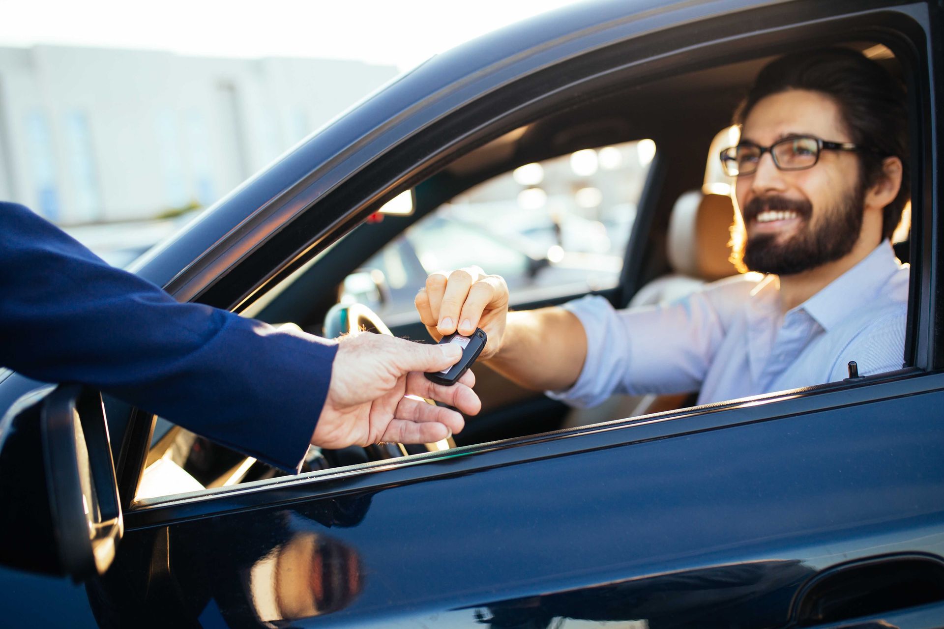 A person hands a car key fob to a smiling individual sitting in the driver's seat of a dark vehicle.