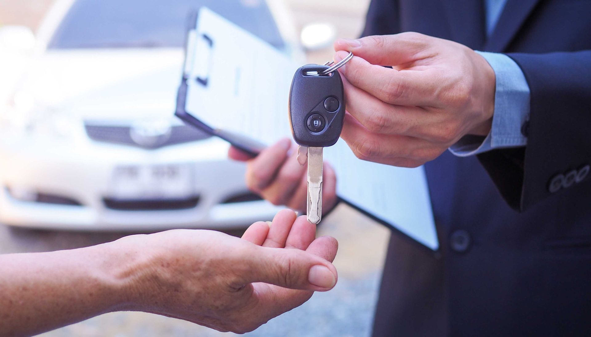 Une personne en costume remet des clés de voiture à une autre, avec une voiture et un bloc-notes flous en arrière-plan.