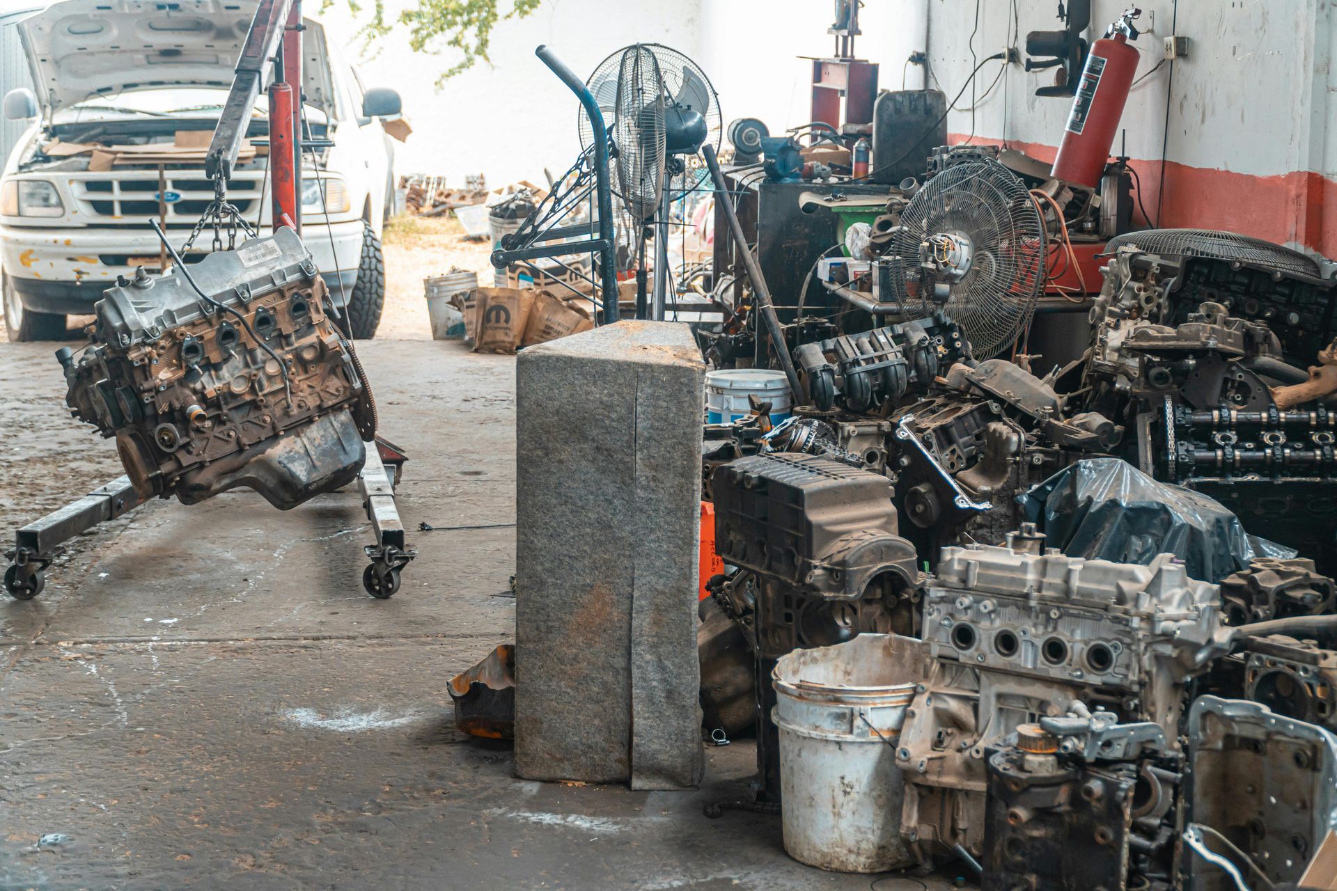 Un atelier de réparation automobile en désordre, avec un moteur sur un support, un camion garé et des piles de pièces de moteur diverses sur le sol en béton.