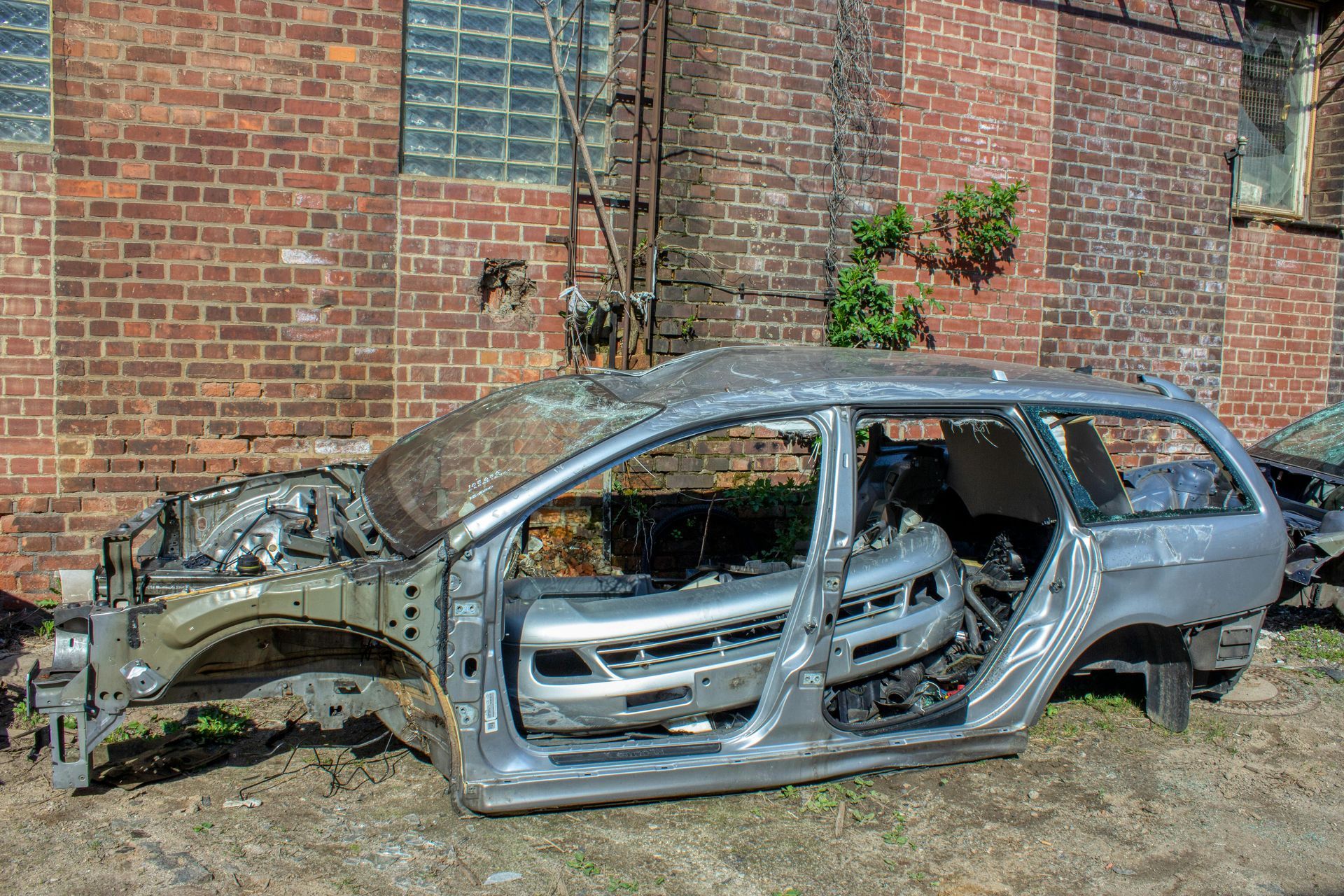 Une carrosserie argentée dépouillée est garée à l'extérieur, adossée à un mur de briques brunes, avec une échelle et de la végétation verte.