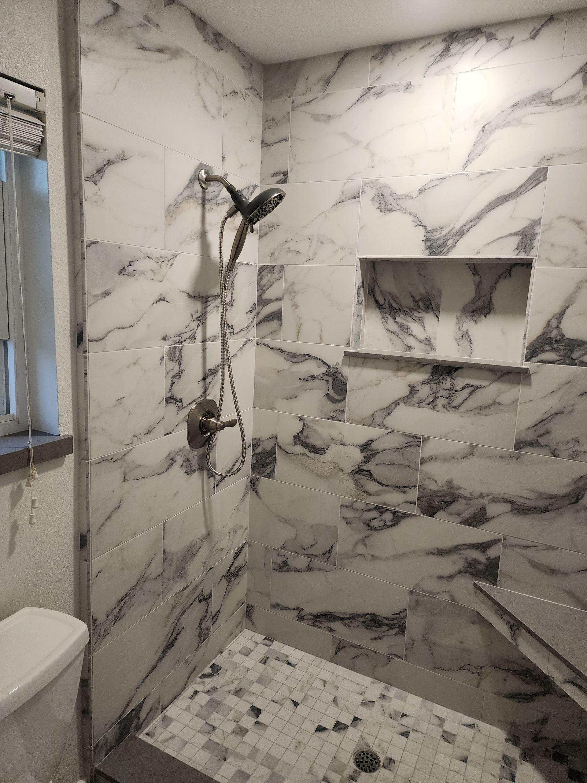 Marble-tiled shower with mosaic floor, built-in shelf, and black shower fixtures