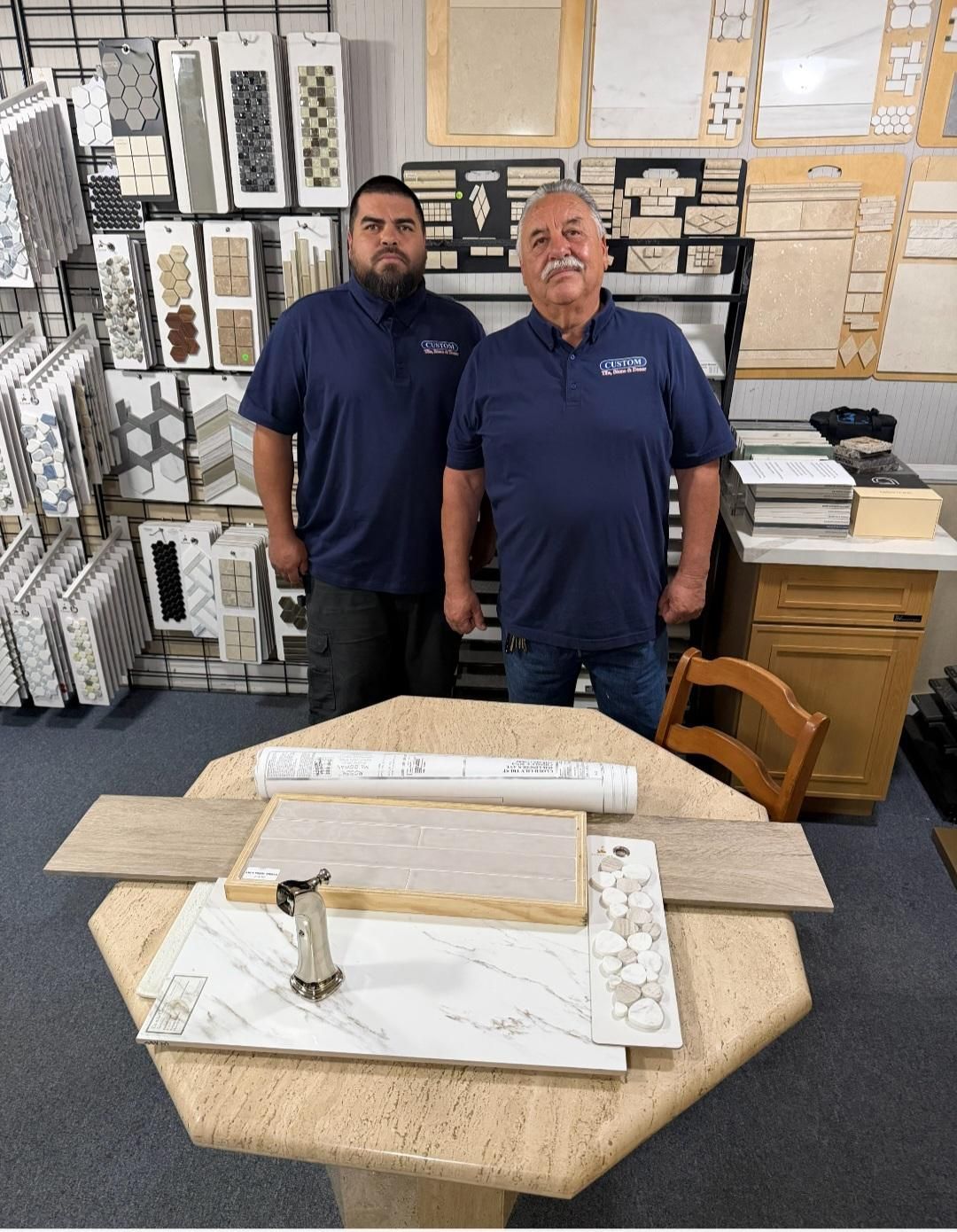 Two men in navy shirts standing in a hardware store behind a display table with tile samples and tools