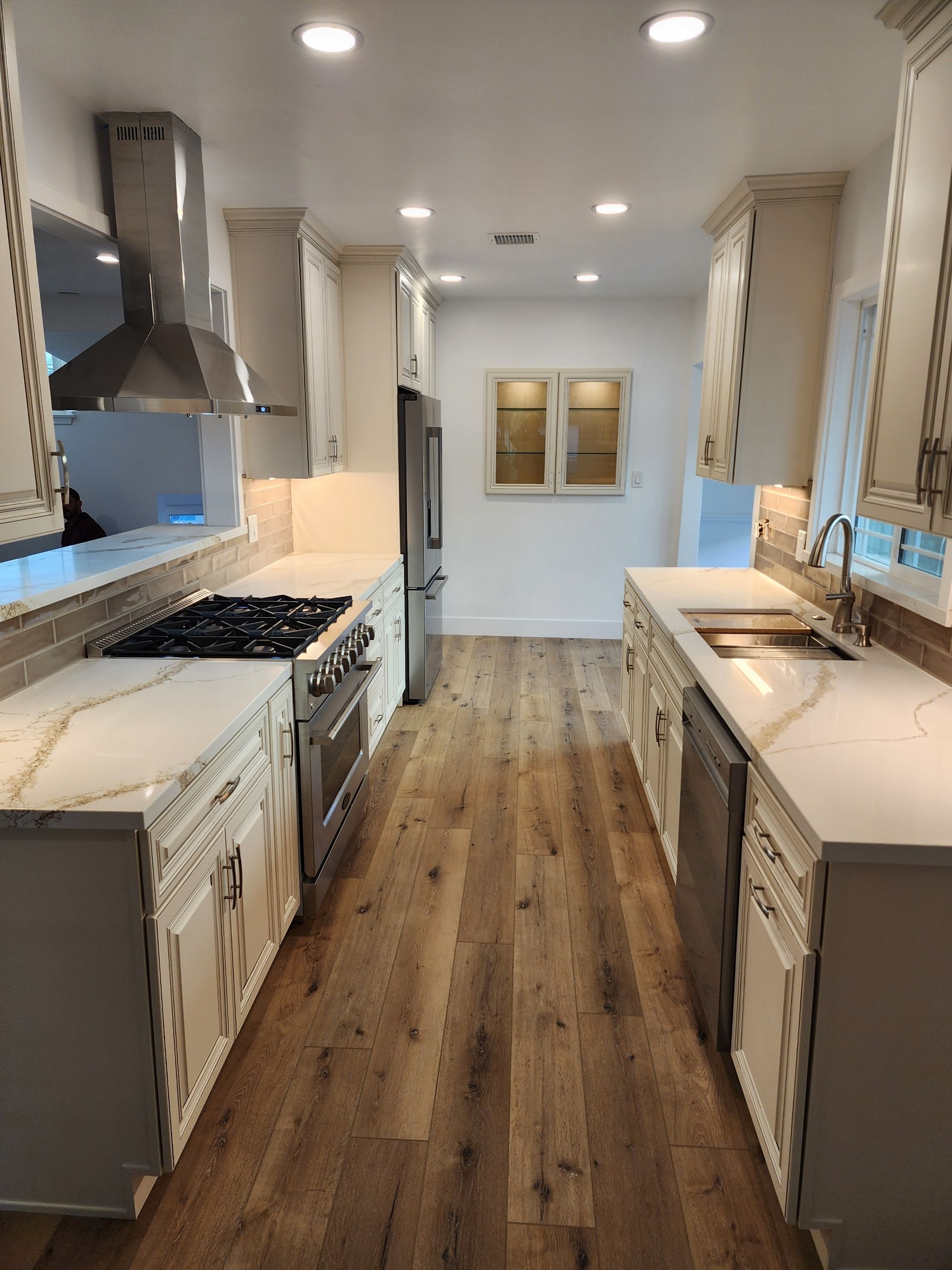 Bright narrow kitchen with white cabinets, wood floor, stainless range, and a long central walkway