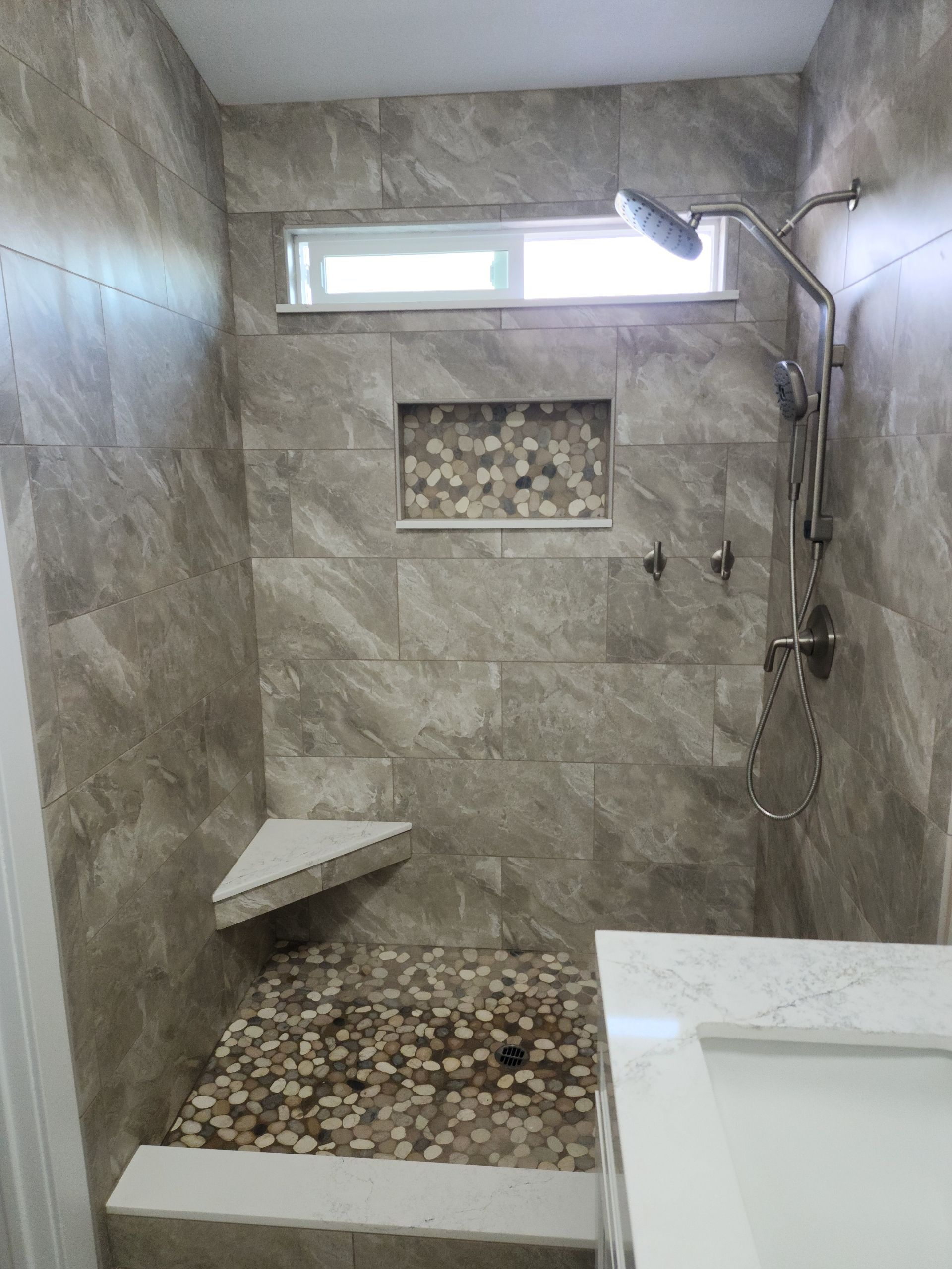 Small tiled walk-in shower with pebble floor, built-in shelf, and handheld showerhead.