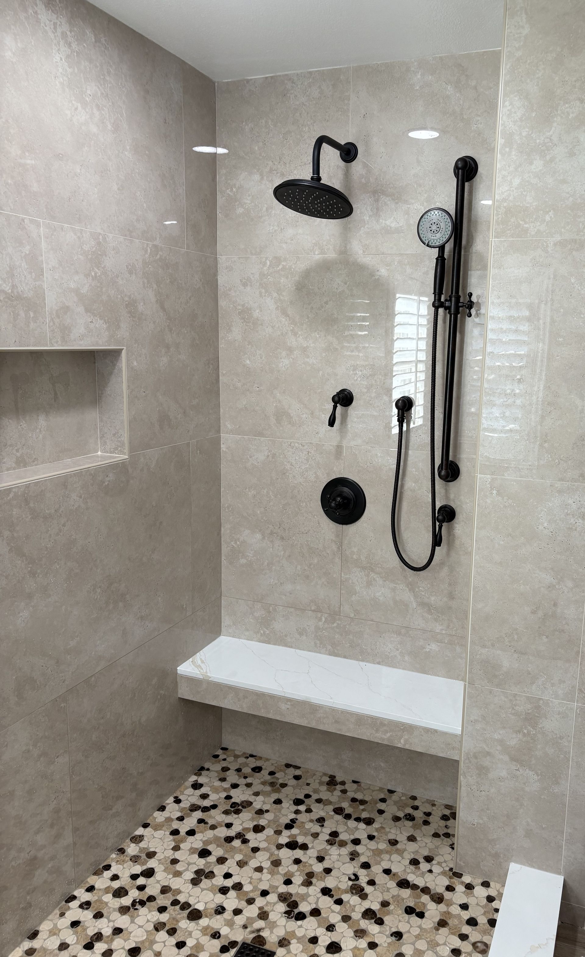 Modern walk-in shower with gray tile, black fixtures, built-in bench, and pebble mosaic floor.