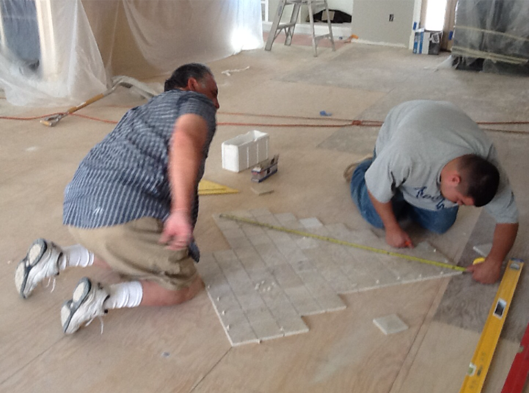 Two workers kneel on a concrete floor, measuring and marking a surface with tools.