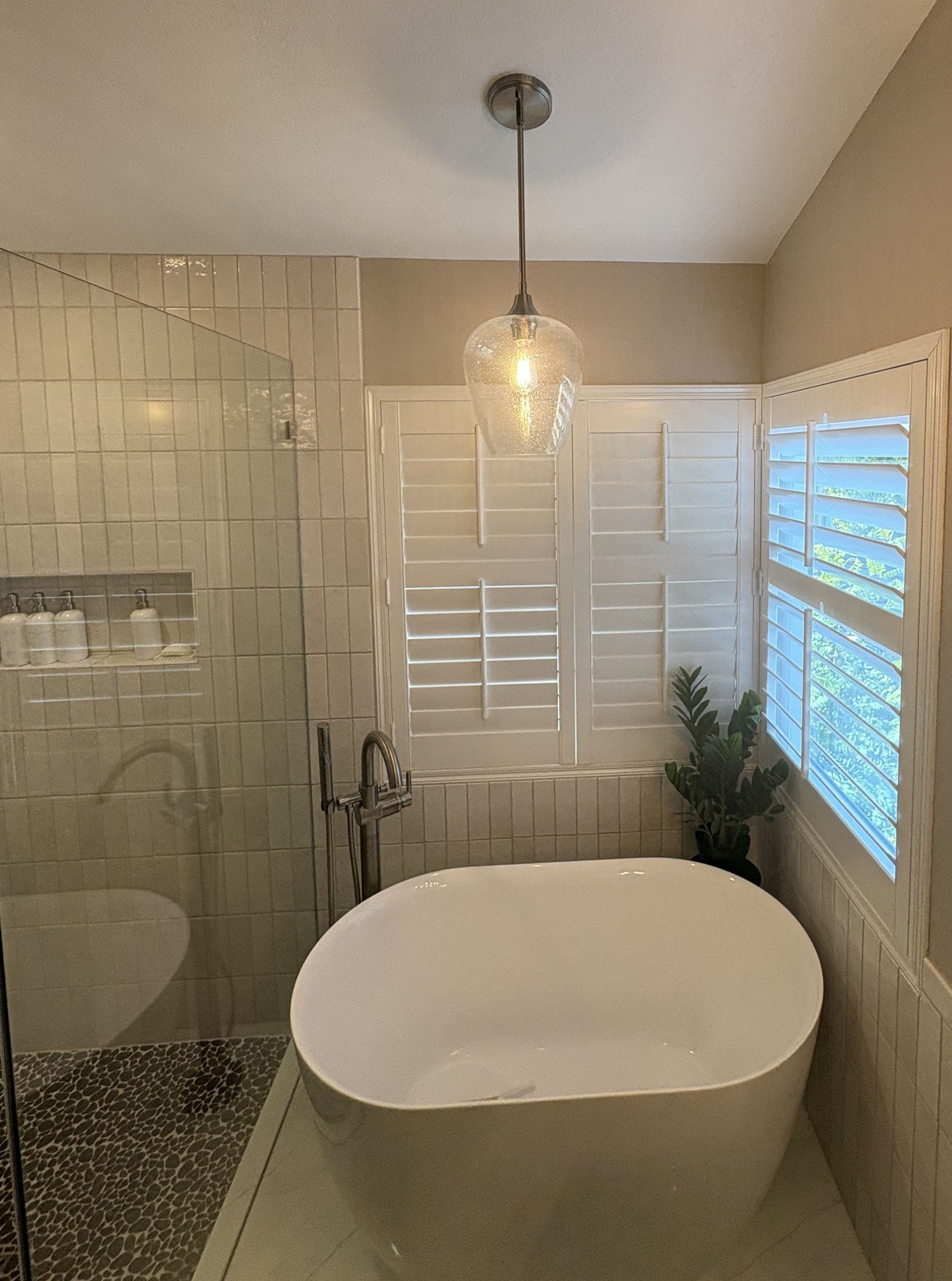 Modern bathroom with a freestanding tub, glass shower, shuttered window, and pendant light.