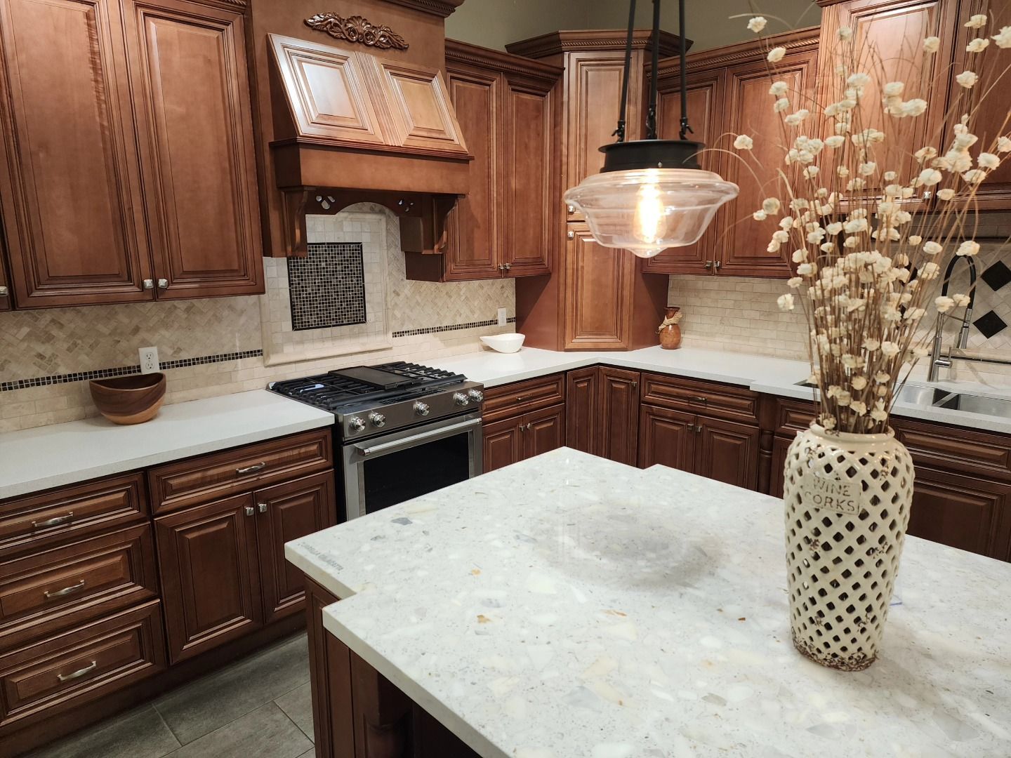 Warm kitchen with dark wood cabinets, white countertops, and a marble-topped island with a vase of dried flowers.