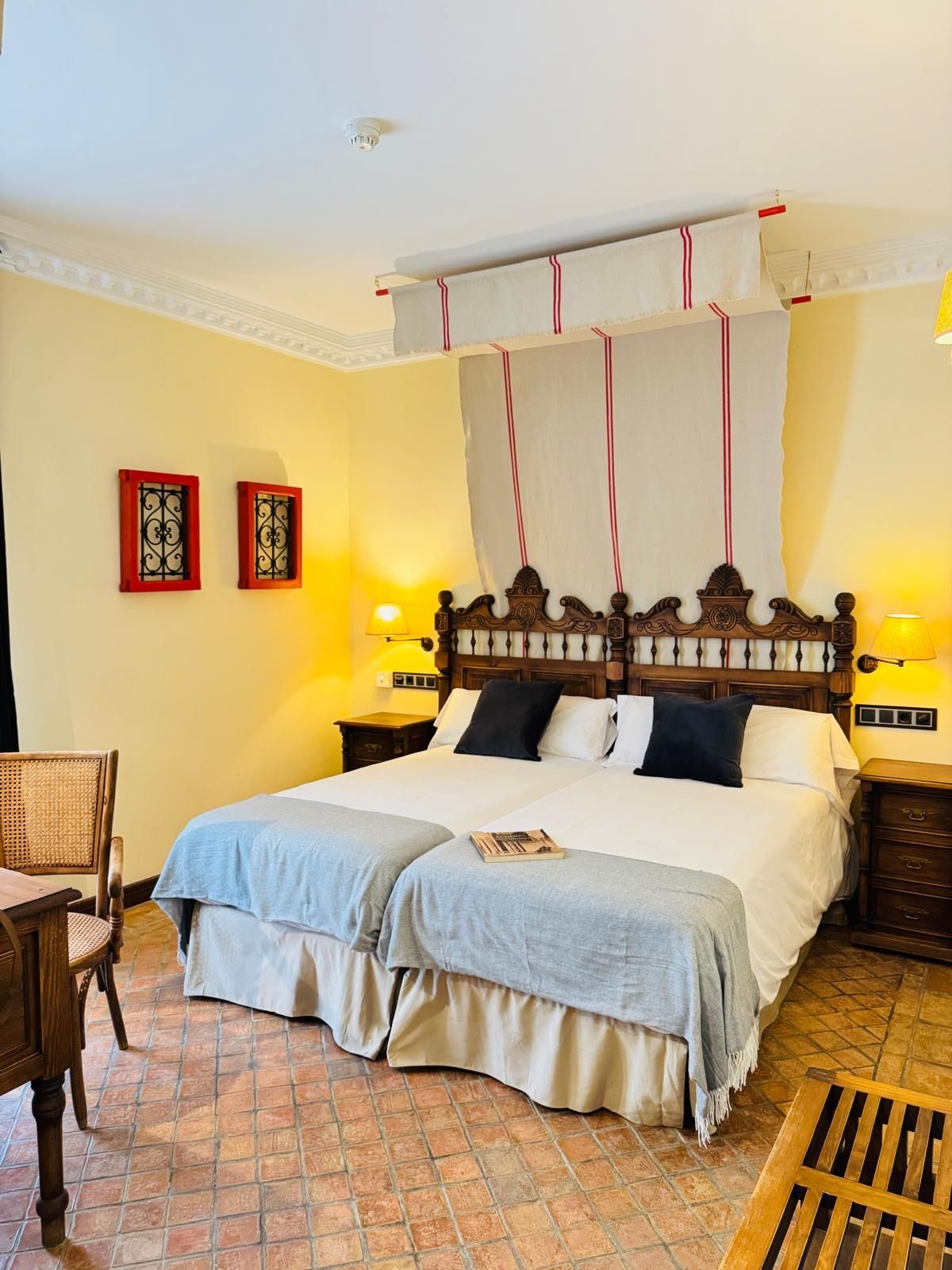 Habitación acogedora con cama de madera oscura tallada, ropa de cama blanca y lámparas cálidas en un entorno rústico.