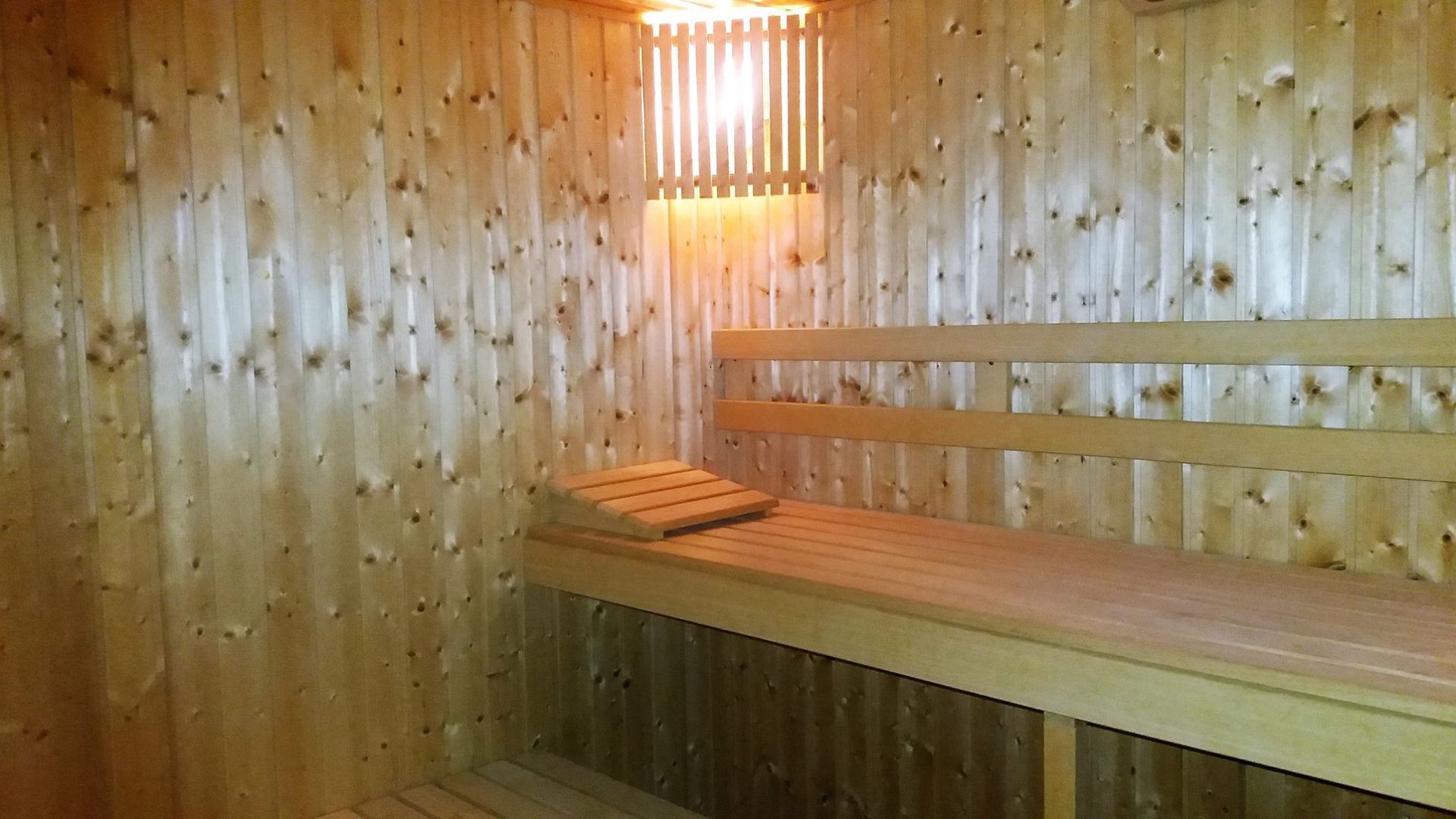 Sauna revestida de madera con un banco de madera y luz cálida proveniente de un calefactor cerca del techo.