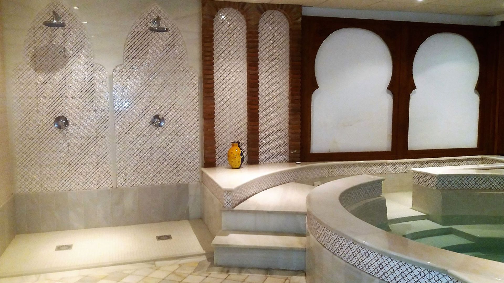 Spa interior revestido de azulejos con ventanas arqueadas, duchas y un borde de piscina curvo.