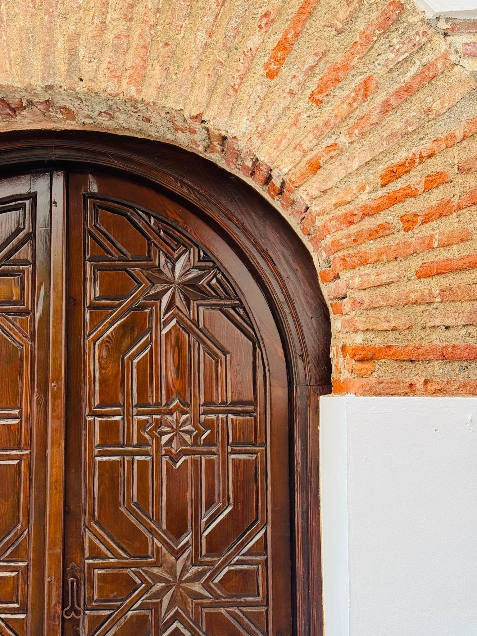 Puerta de madera tallada y ornamentada en un arco de ladrillo con una pared blanca al lado.