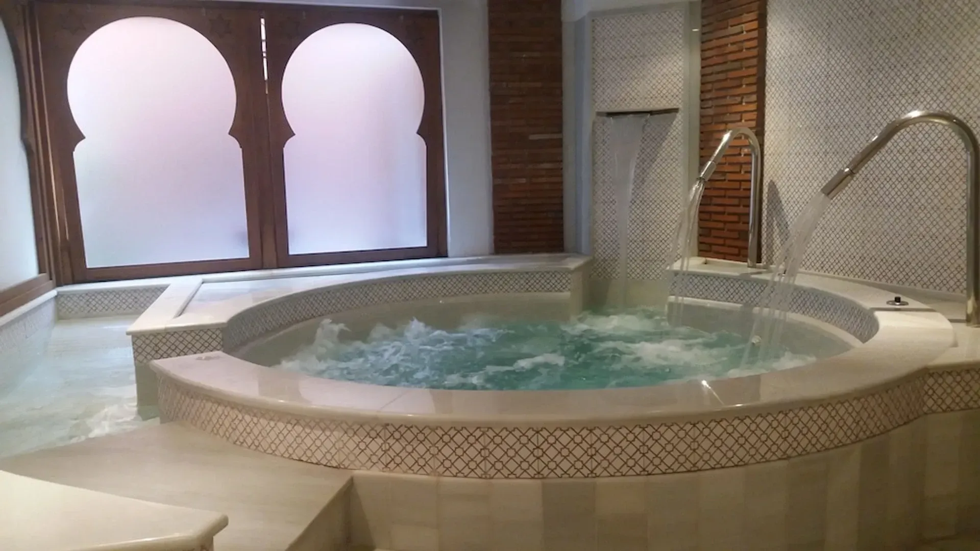 Bañera de hidromasaje interior con revestimiento de azulejos, ventanas arqueadas y grifo cromado en un baño tipo spa.
