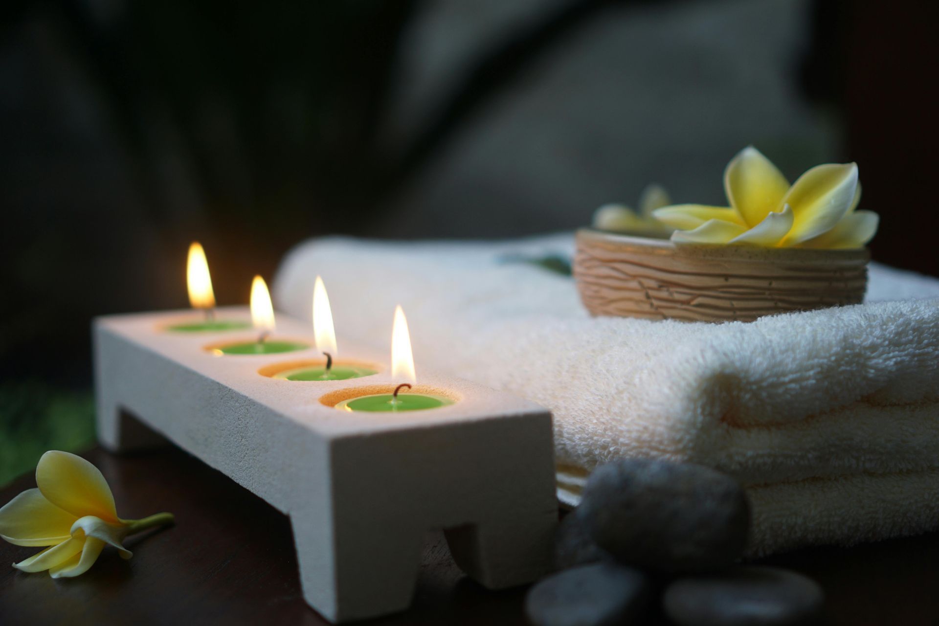 Velas de spa junto a una toalla blanca doblada, piedras y flores amarillas en un entorno tranquilo.