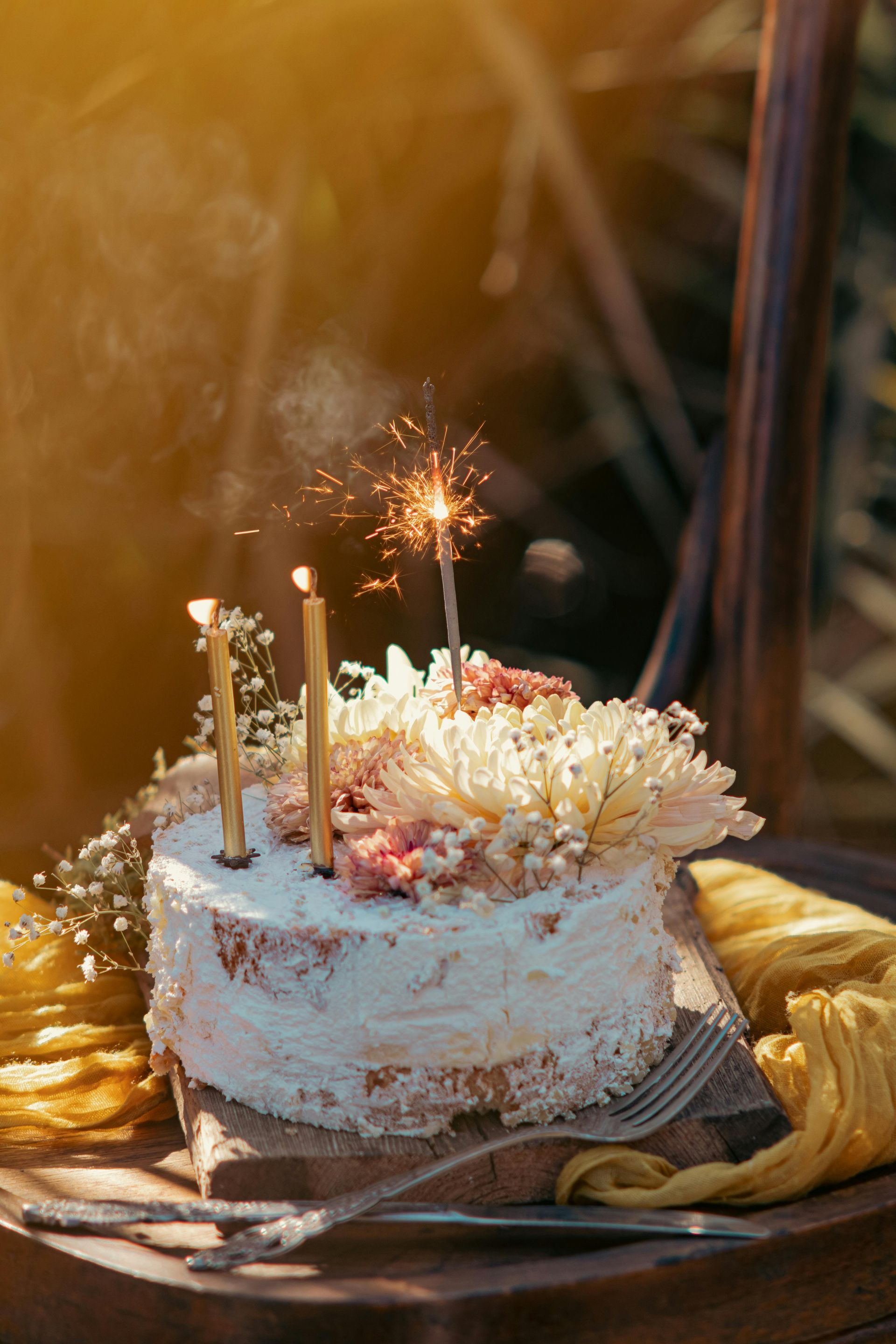 Tarta de cumpleaños con velas encendidas, glaseado blanco y flores sobre una mesa.