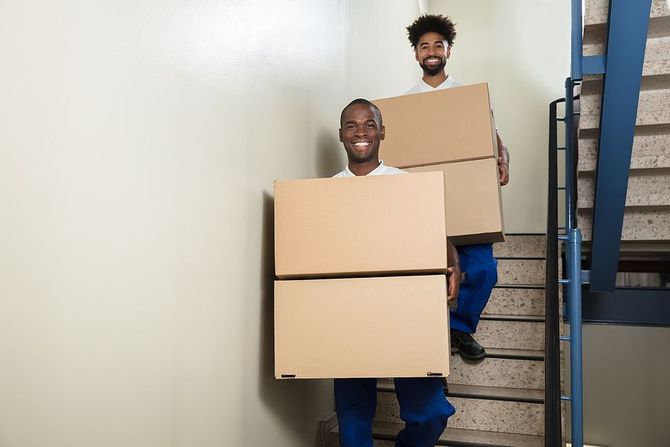 Two smiling movers carrying stacked moving boxes up a staircase.