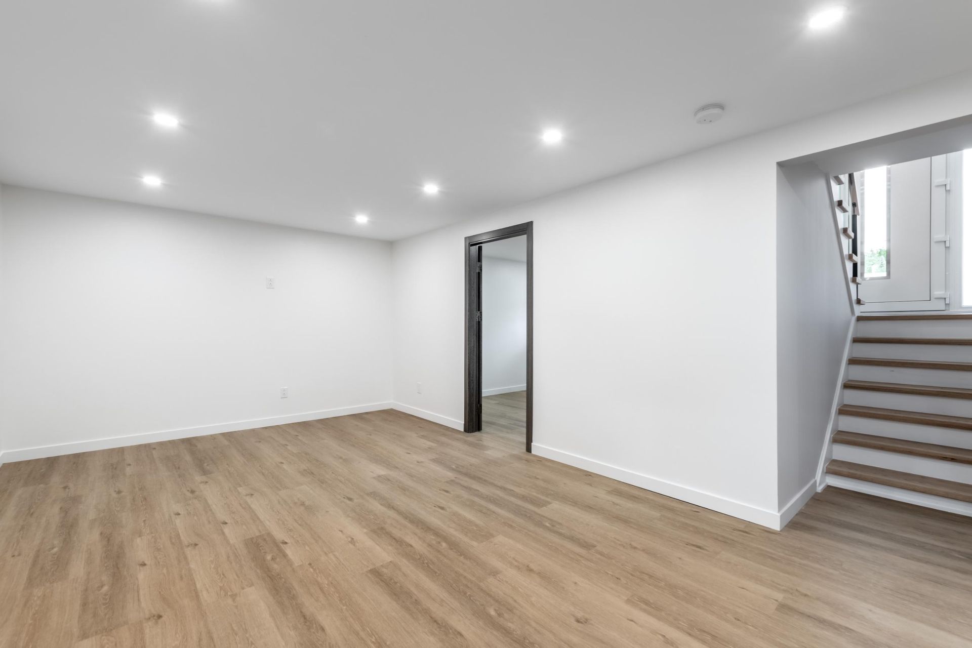 Empty room with light wood flooring, white walls, recessed lighting, and a staircase.