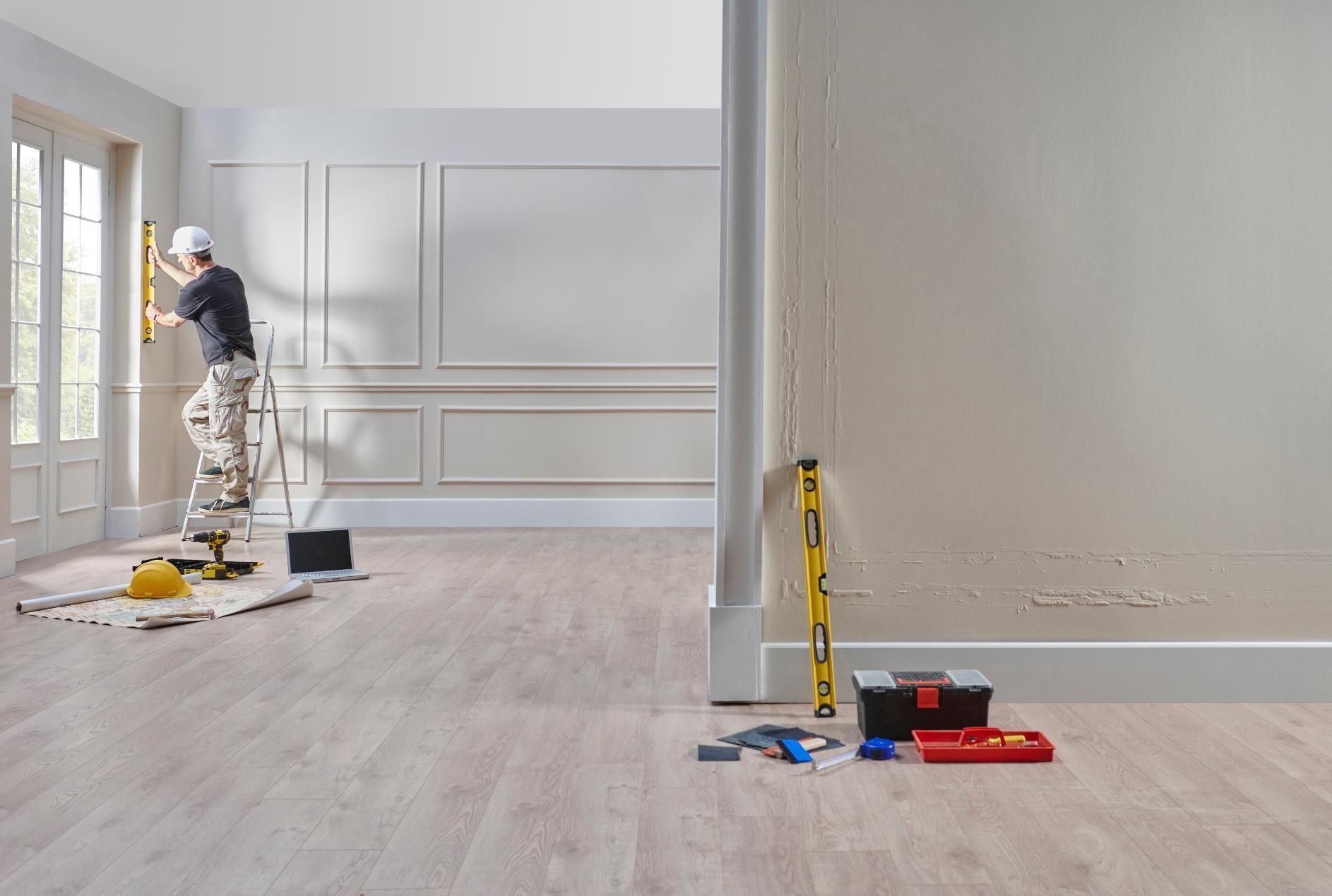 Person on ladder uses level in a room under renovation. Tools on floor. Light wood flooring.