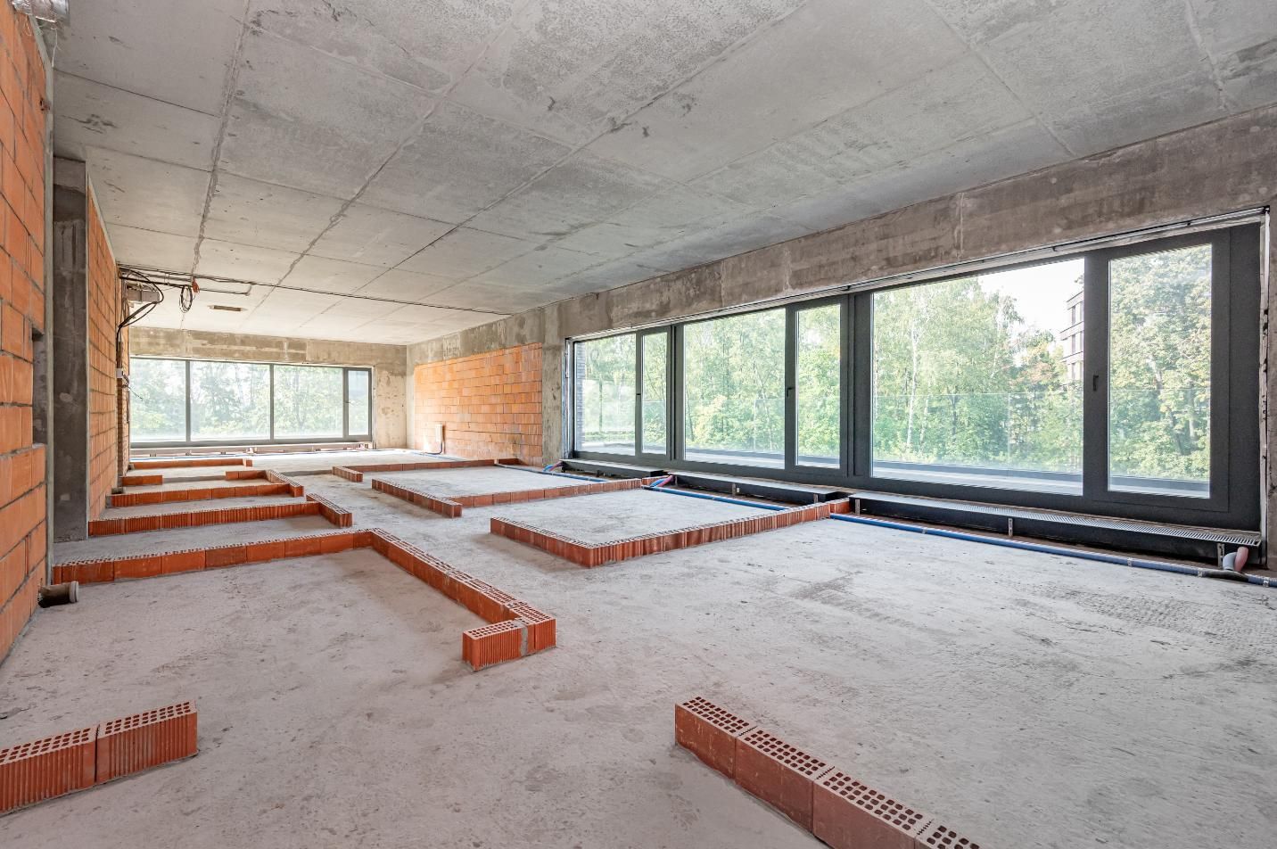 Interior of a building under construction. Unfinished walls, large windows, and brickwork on floor. Concrete ceiling.