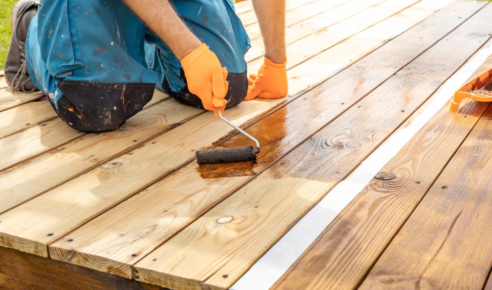 Person applying wood stain to a wooden deck with a roller, wearing orange gloves.