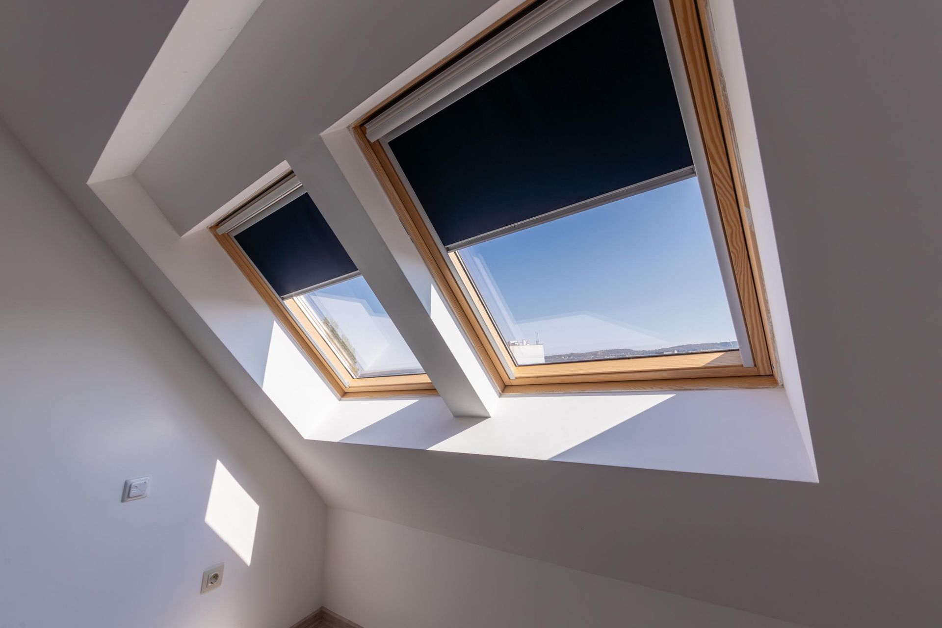 Two angled skylight windows with dark blue shades in a white room, letting in sunlight.