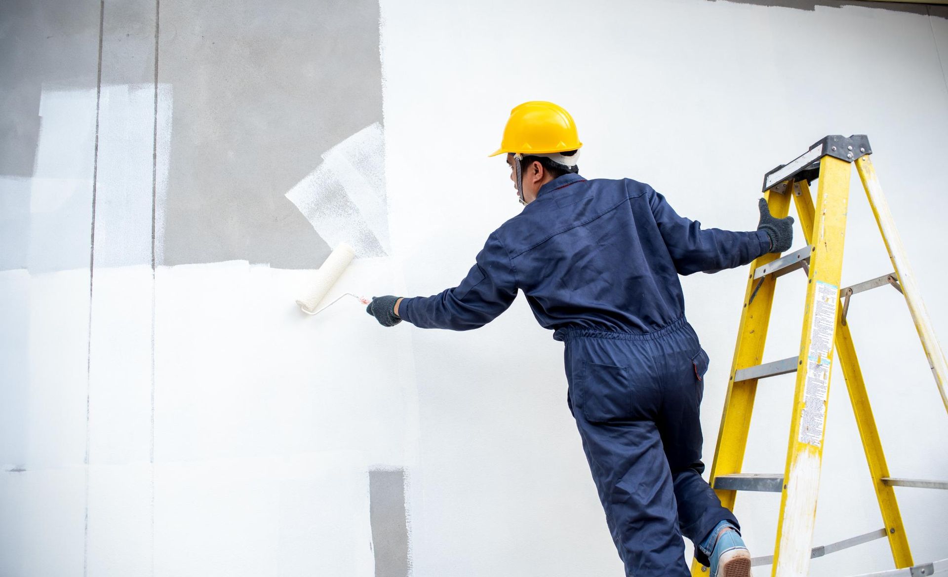 Person in blue jumpsuit and yellow hard hat paints a wall with a roller.