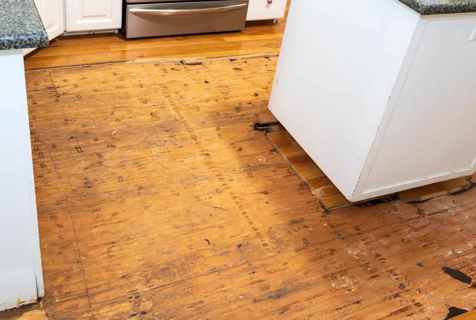Kitchen floor stripped of covering, showing damaged wood planks. White cabinets and stainless steel appliance are visible.