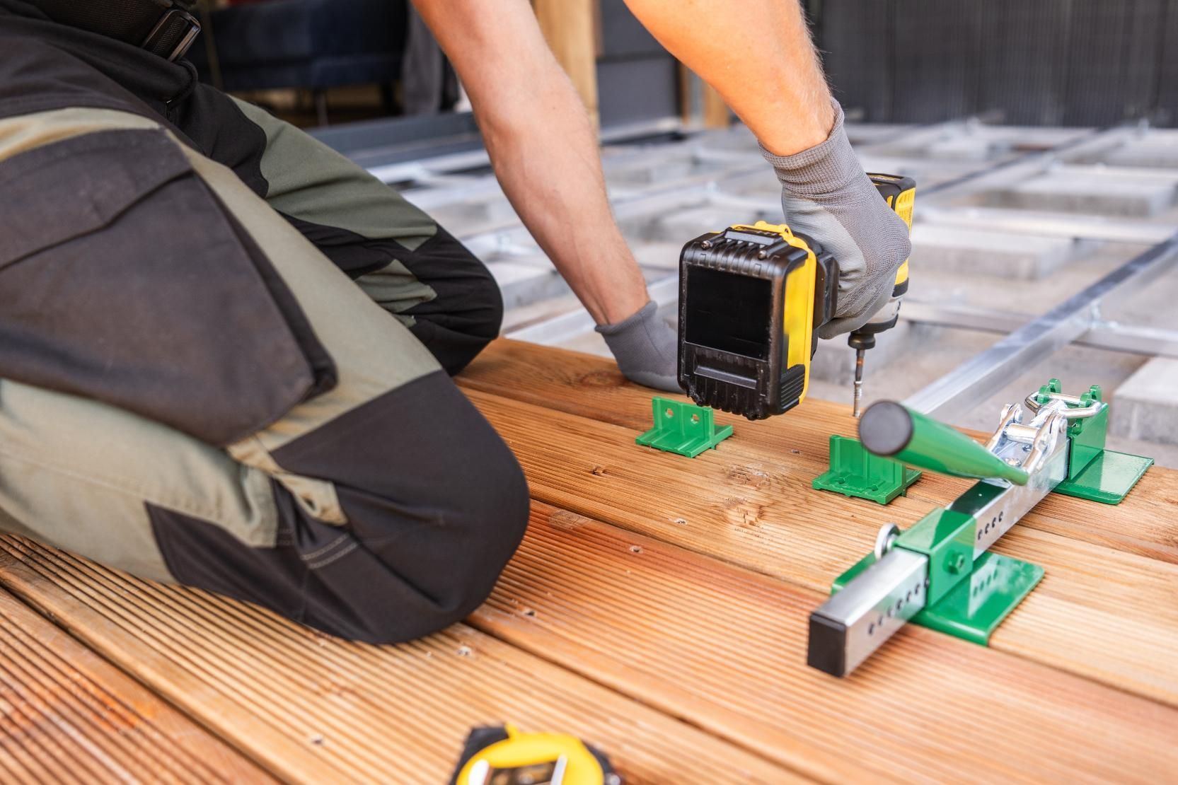 Person using a drill to install wooden deck boards, with green spacers and a measuring tape.