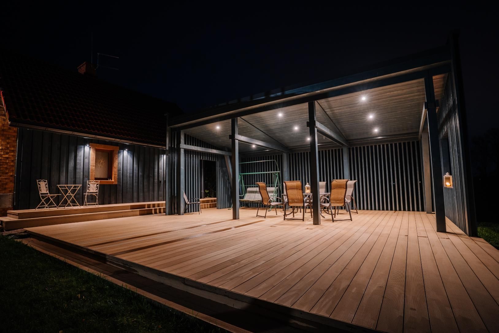 Night view of a wooden patio with overhead lighting, outdoor dining furniture, and a dark building.
