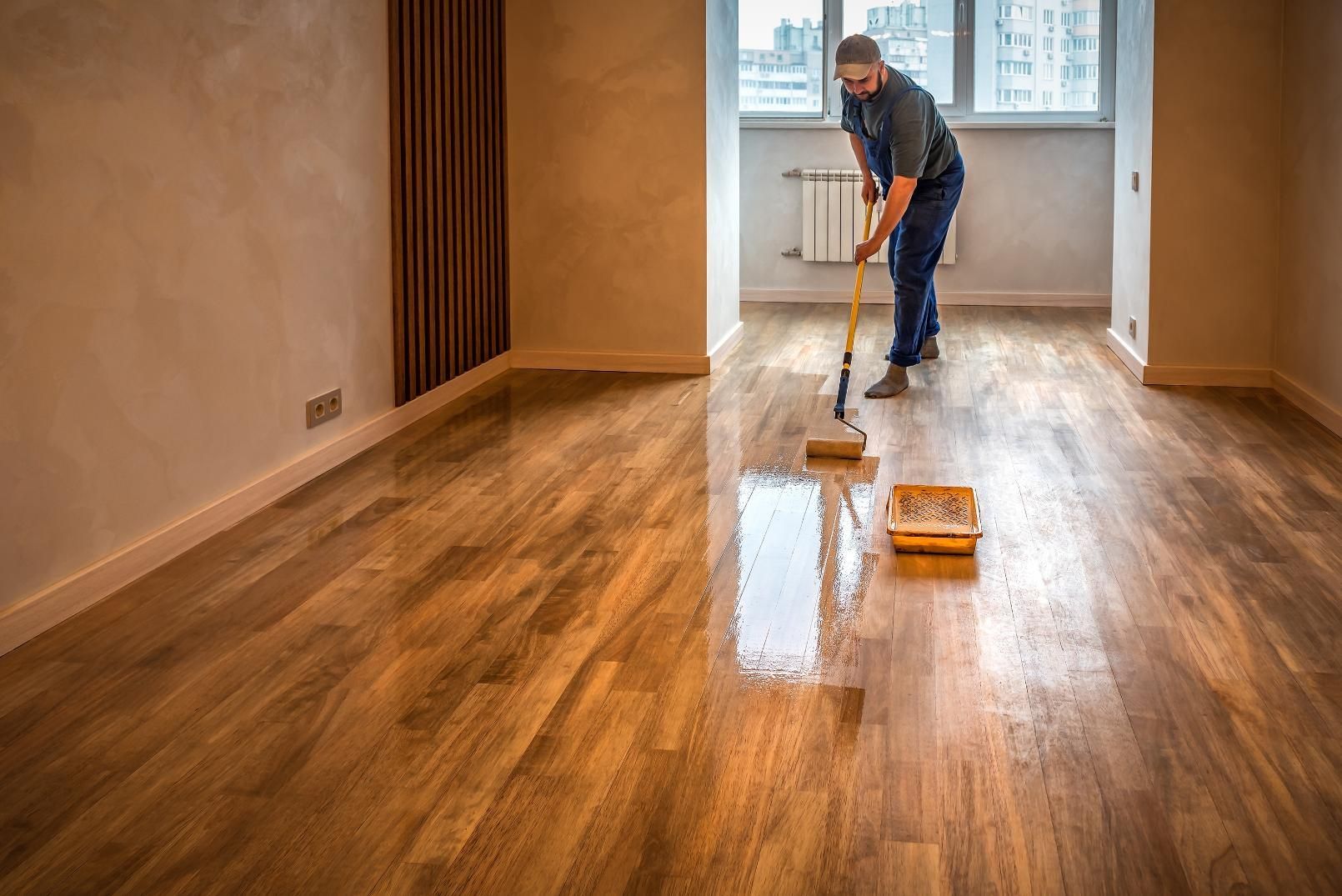 Person applying finish to a wood floor with a roller and tray in an empty room with a window.