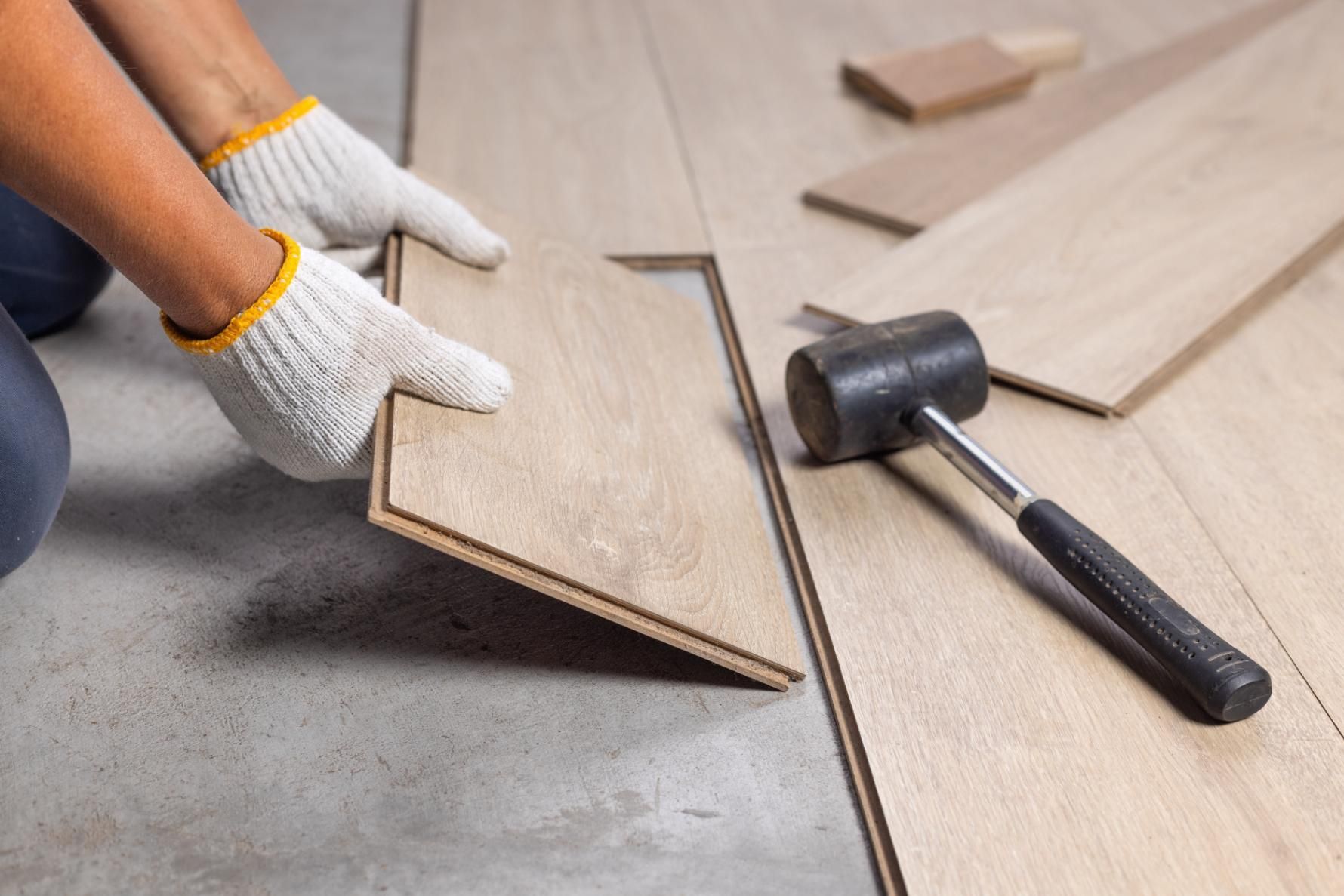 Person installing wood flooring, wearing gloves, with a hammer nearby.