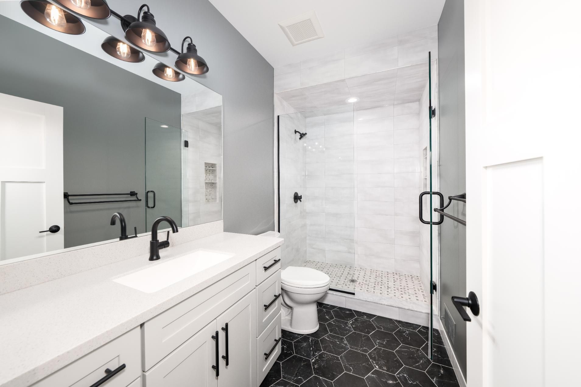 Modern bathroom with white vanity, gray walls, glass shower, black hexagon tile floor.