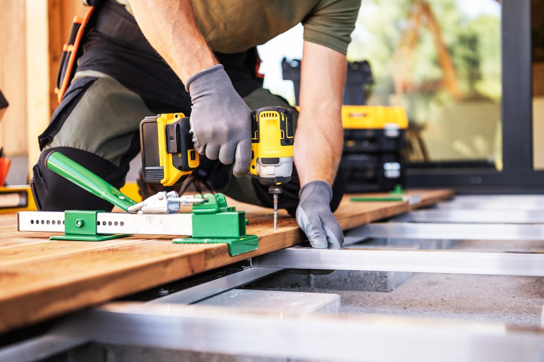 Person using a yellow power drill to fasten a wood plank onto a deck frame.