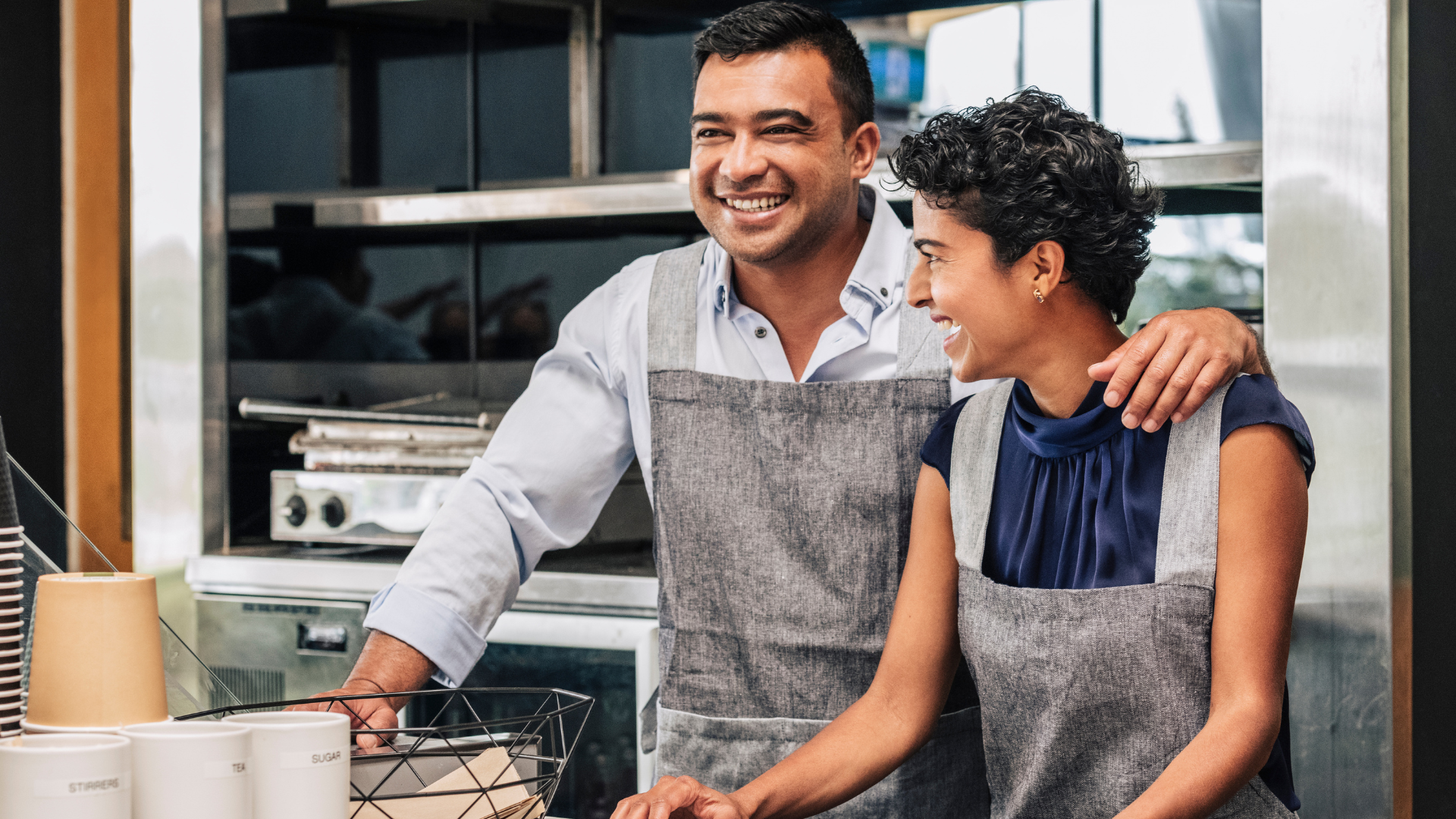 Two people in aprons smiling and cooking together in a bright kitchen