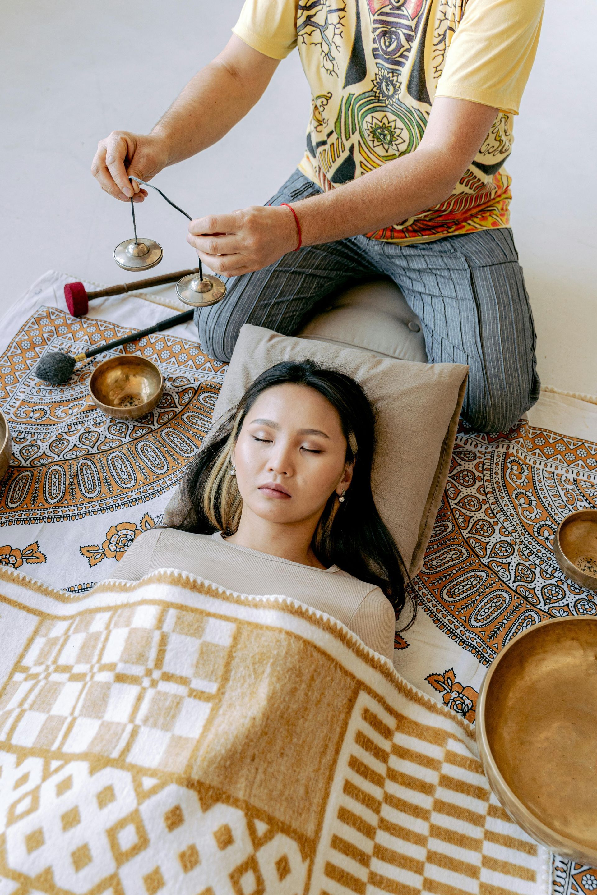 A woman is laying under a blanket while a man plays a singing bowl.
