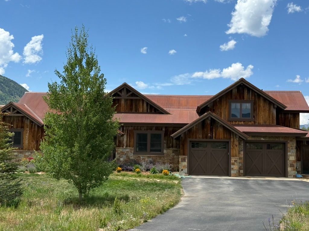 Rustic wooden house with brown roof, garage, and stone accents, on a grassy hill under blue sky.