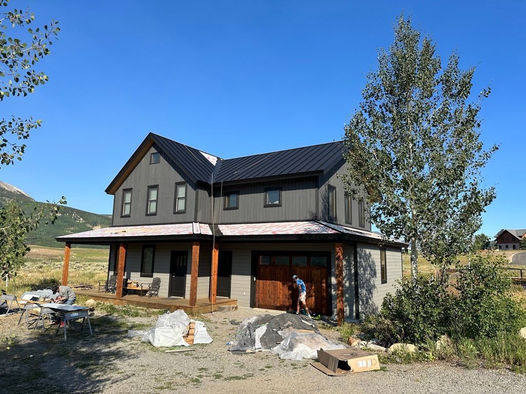 Two-story gray house with black roof, brown garage door, and porch; man working outside.