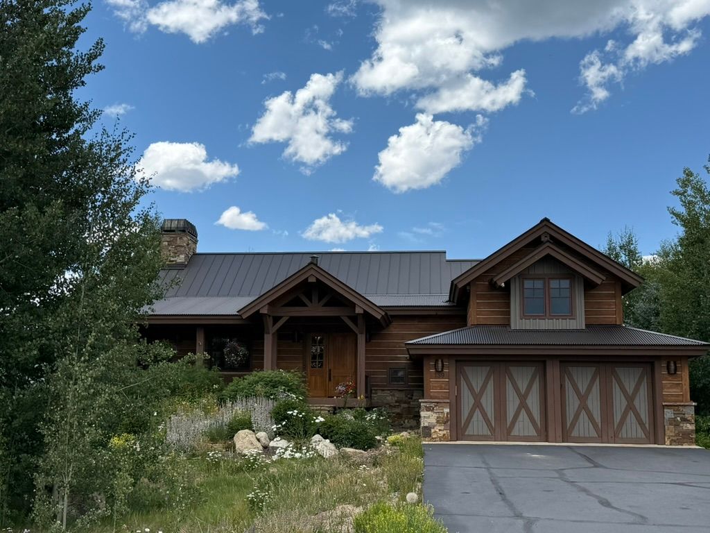 Rustic cabin home with brown wood siding, two-car garage, and black roof under a blue sky with clouds.