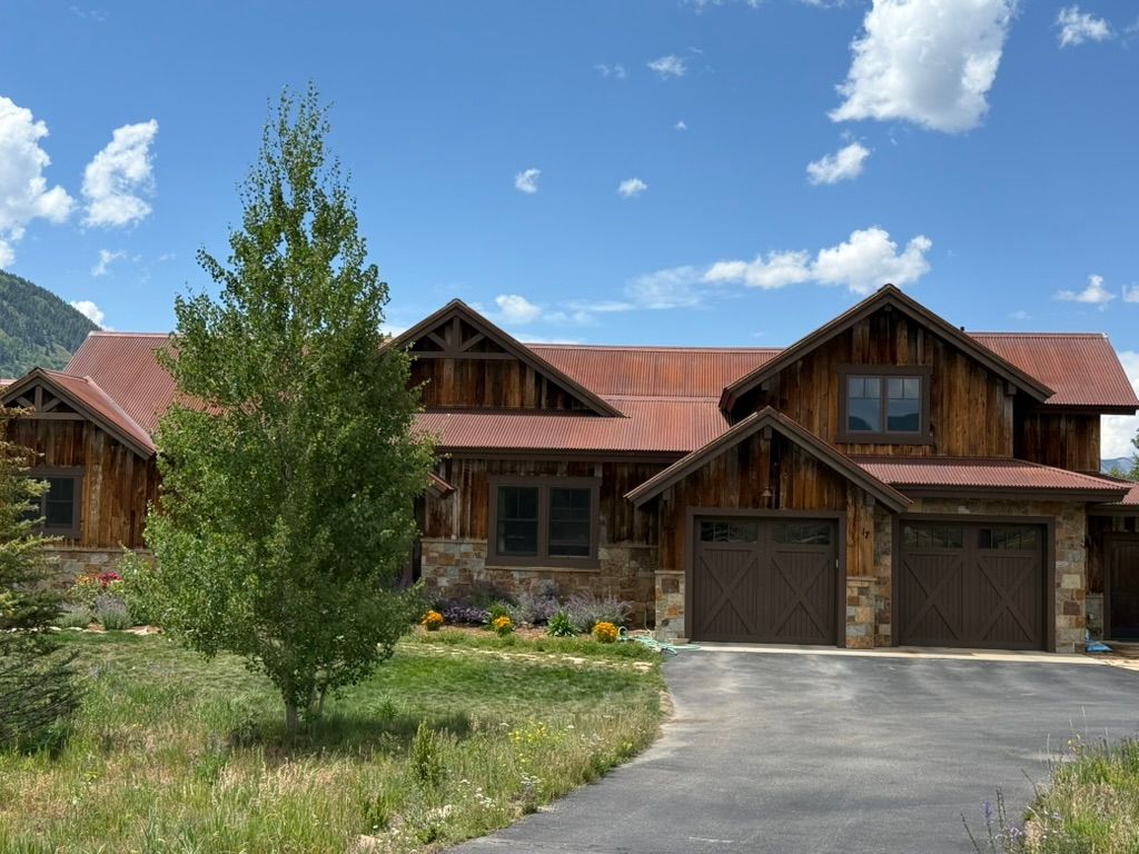Rustic wooden house with a rusty metal roof, surrounded by greenery under a blue sky.