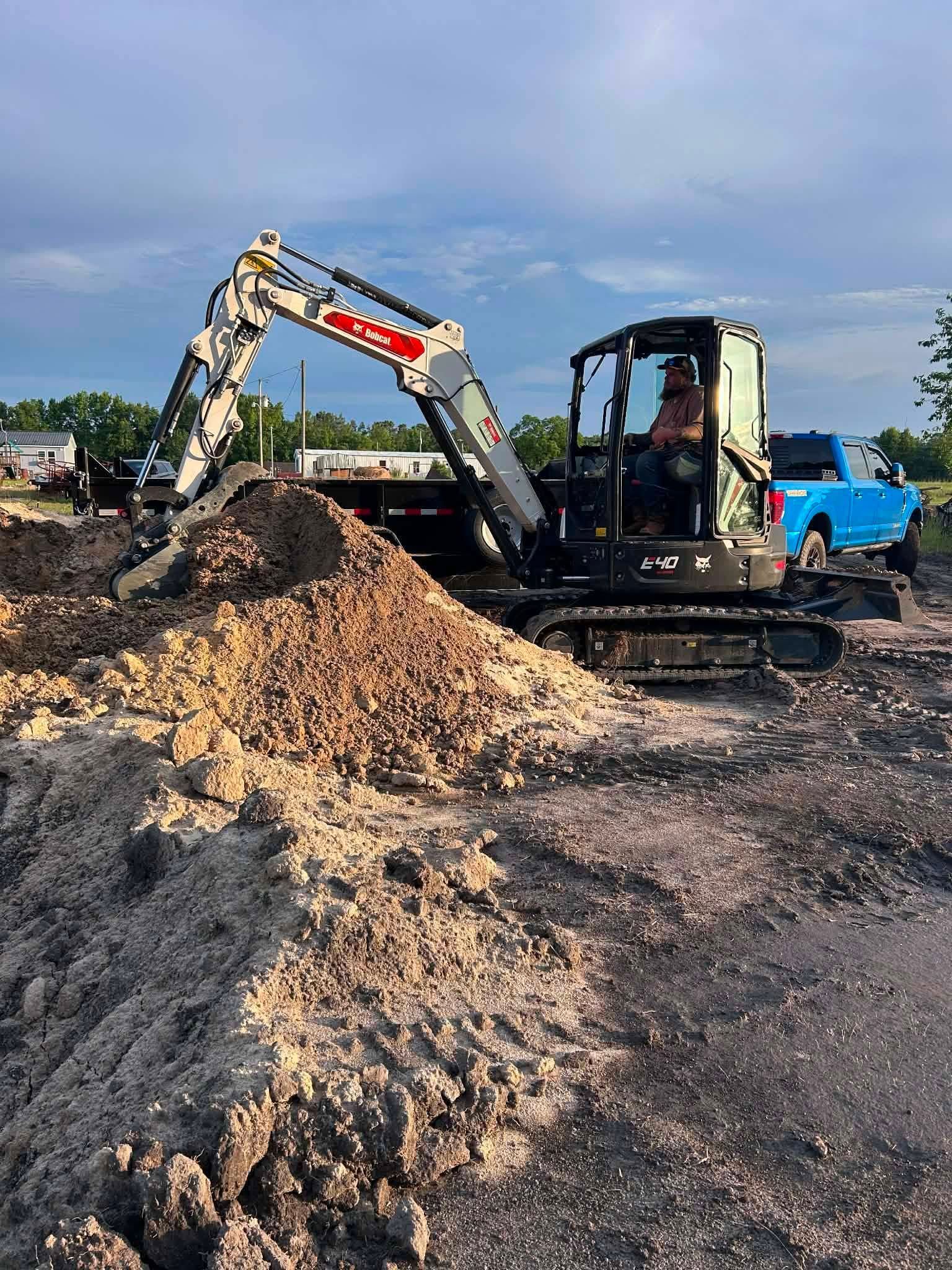 Excavator digging dirt in a construction site beside a blue truck under a cloudy sky