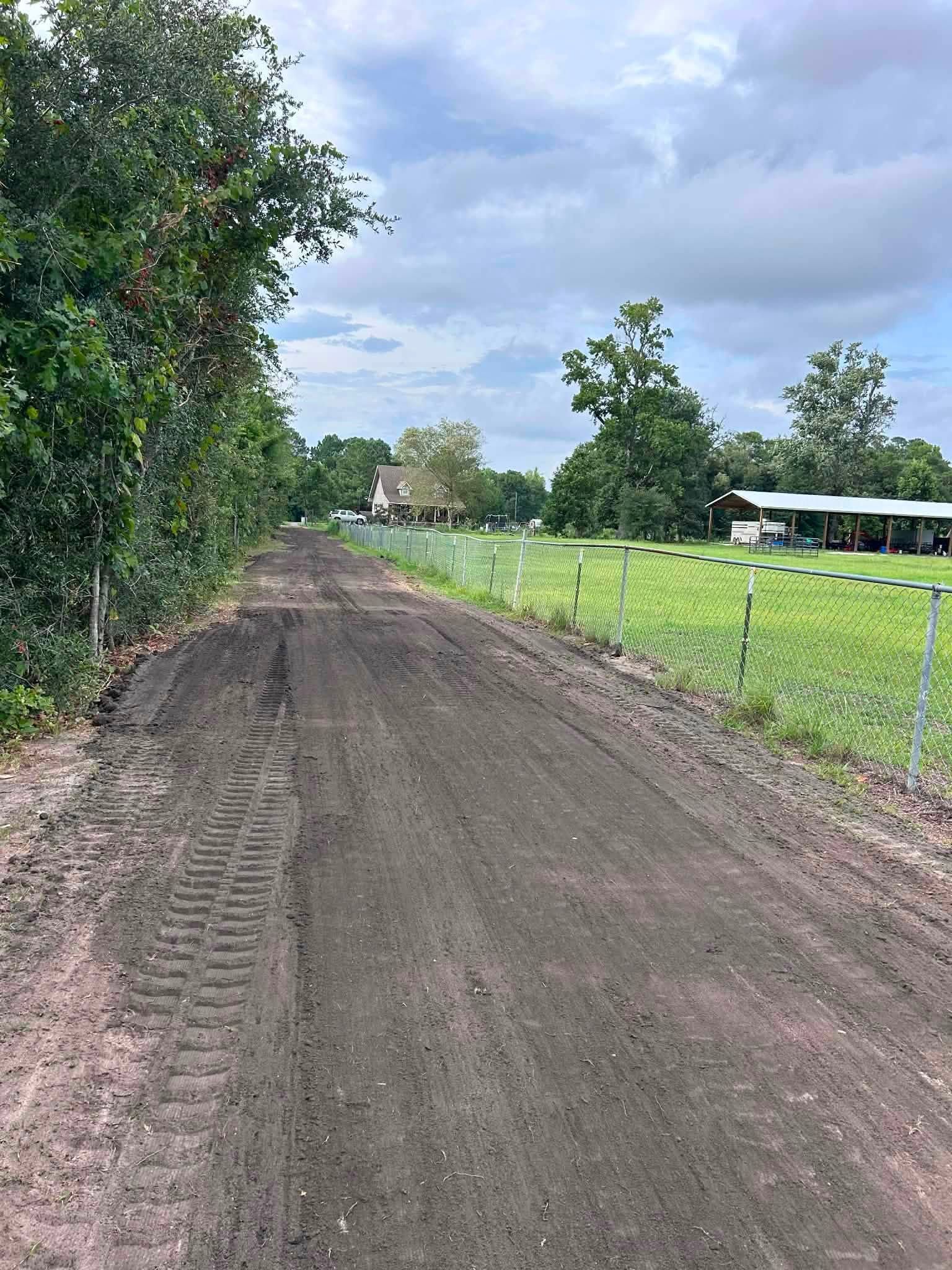 Rural dirt road beside green pasture and trees under a cloudy sky