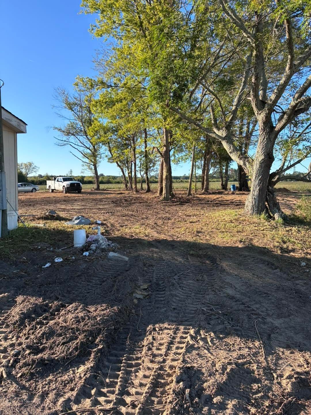 Dirt lot with tire tracks, trees, and a small building under a clear blue sky