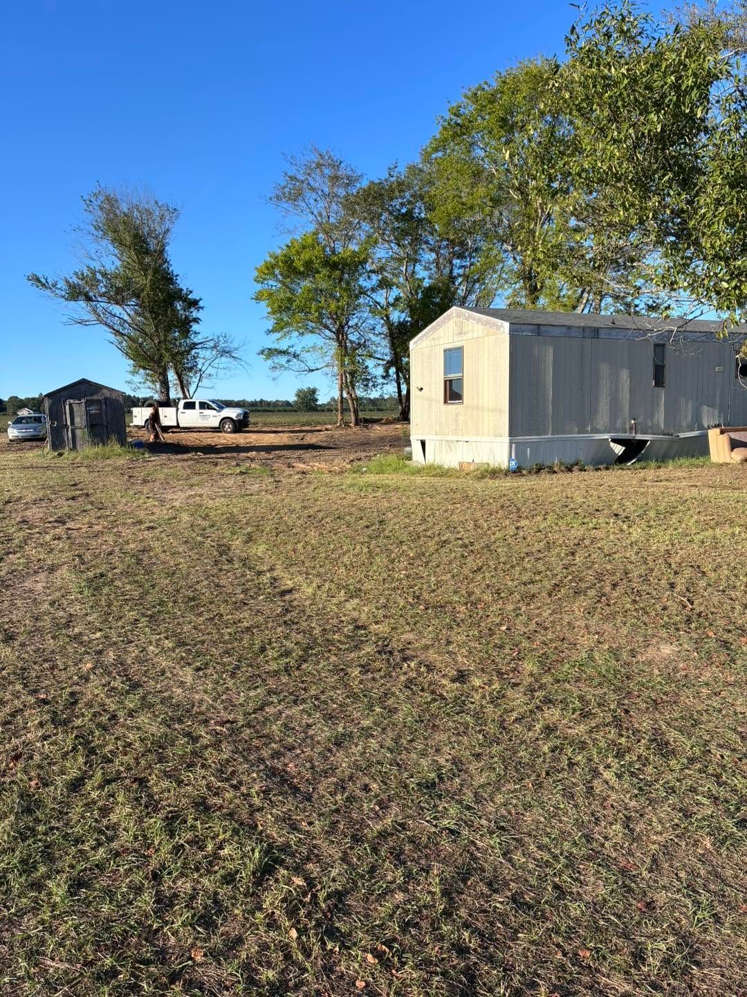 Rural yard with a white mobile home, large trees, and a dirt road under clear blue sky.
